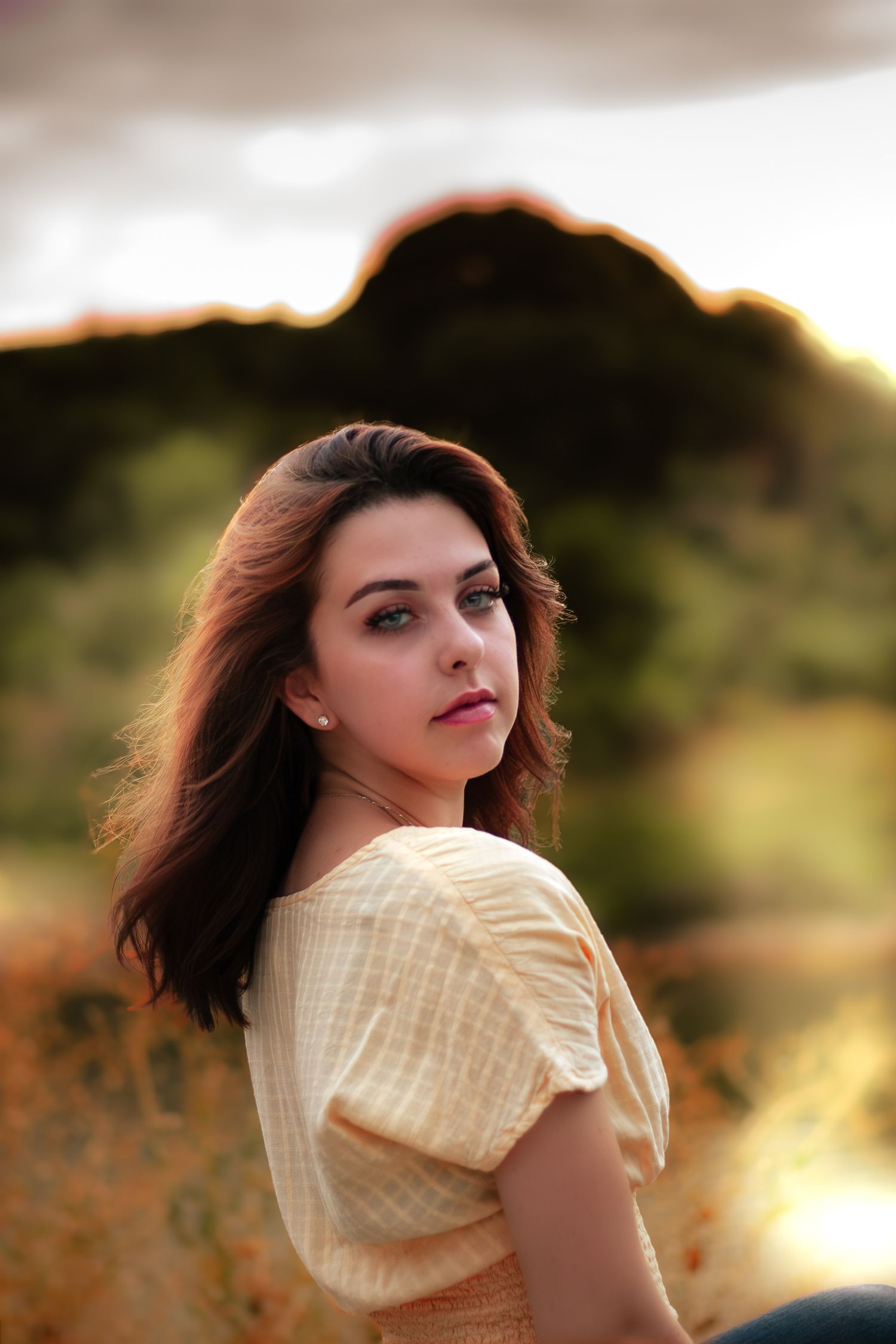 Young woman with auburn hair in yellow top sits in golden field at sunset, gazing at camera.