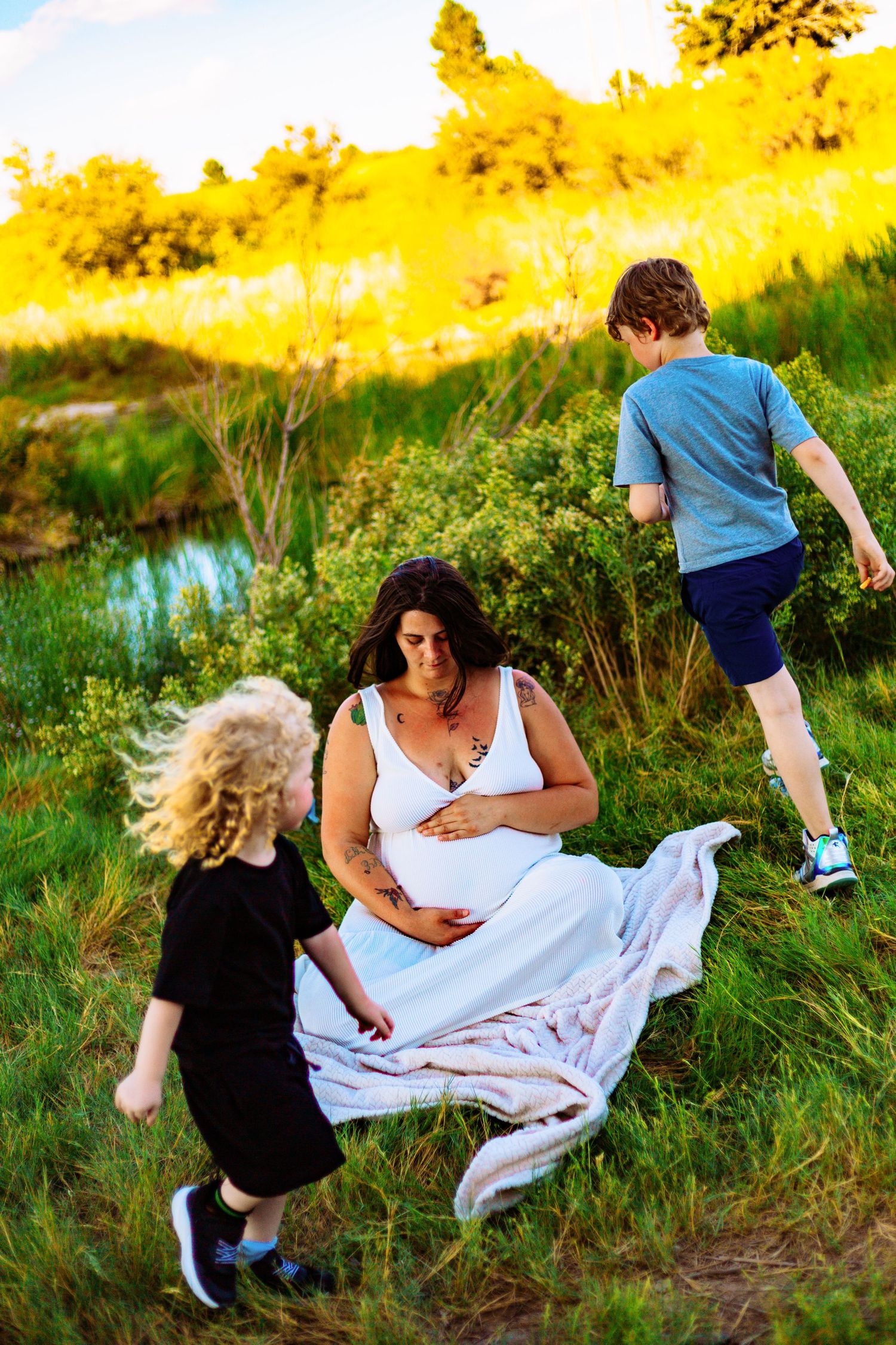 Pregnant woman in white dress sitting on grass with two children playing around her in a sunny green field.