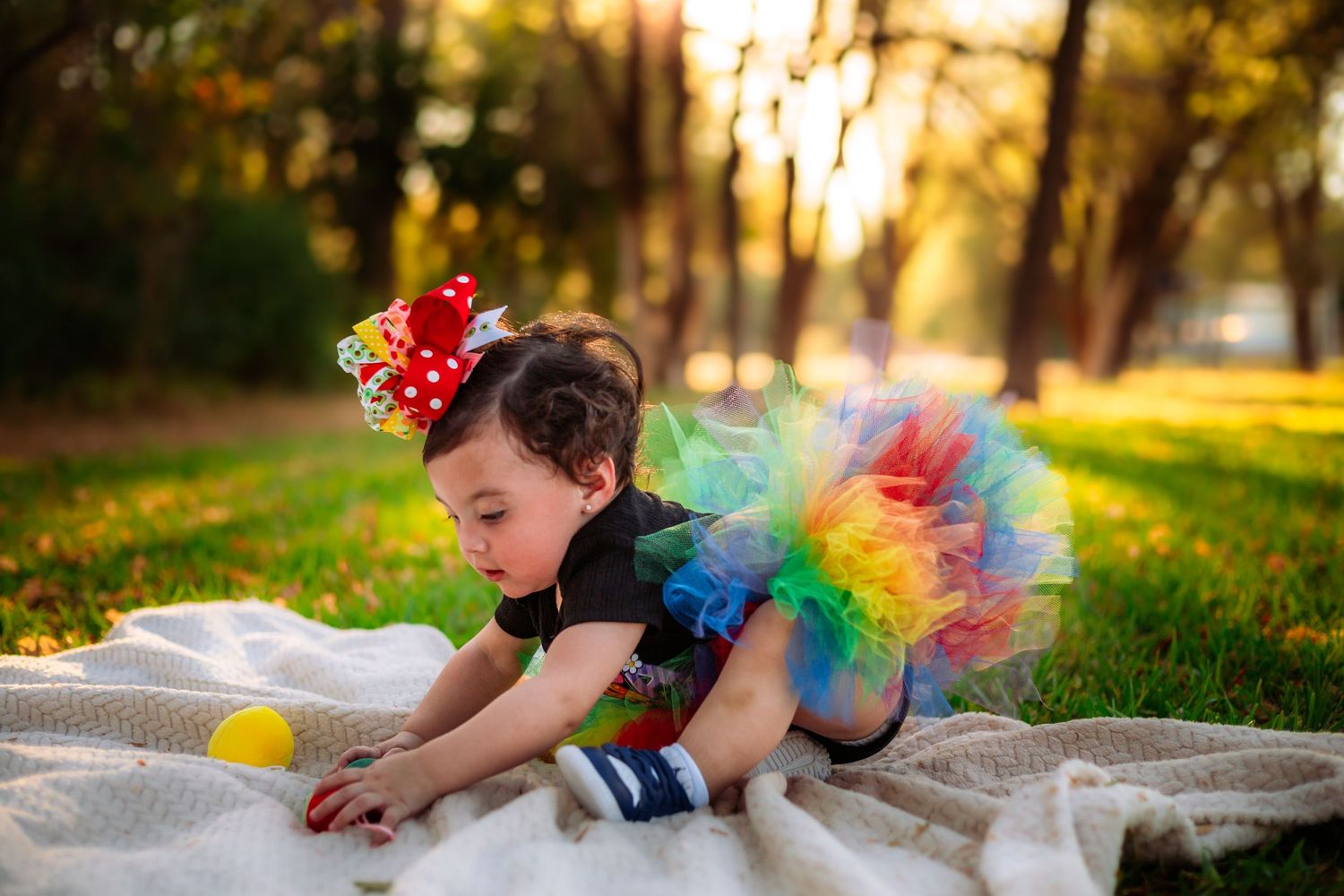Toddler girl in rainbow tutu and polka dot bow crawls on blanket in sunny park setting.