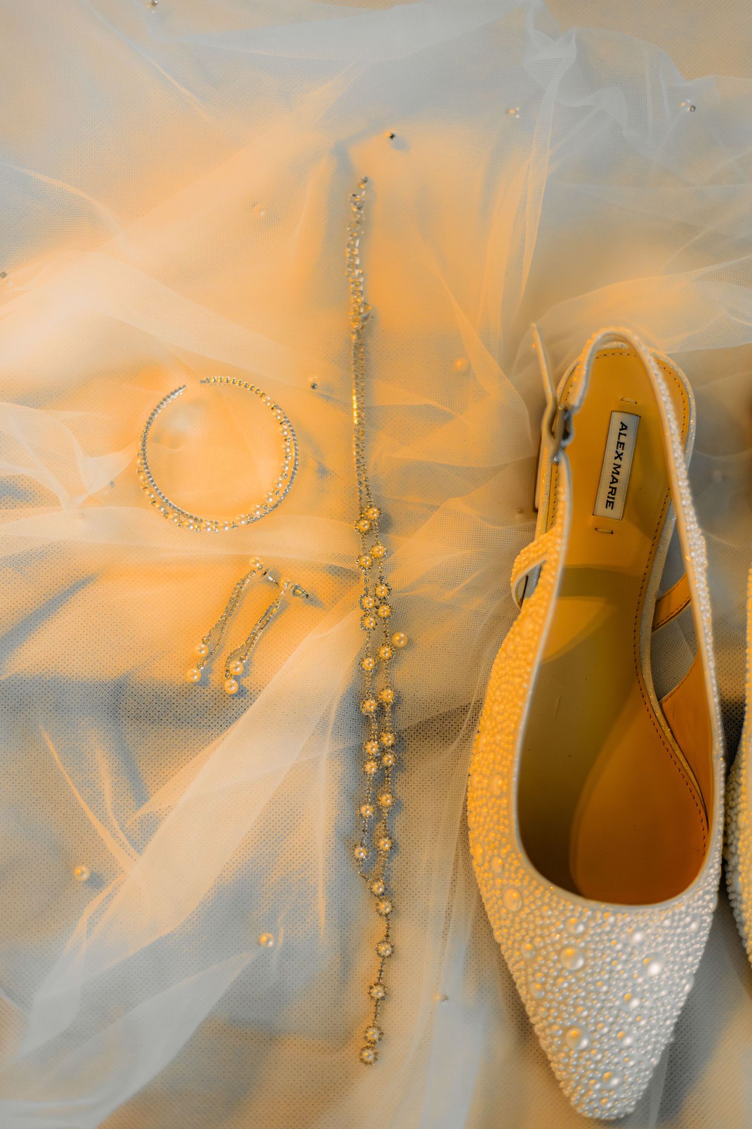 Pearl-embellished bridal slingback heels beside crystal drop earrings and bracelet on a sheer white tulle veil.