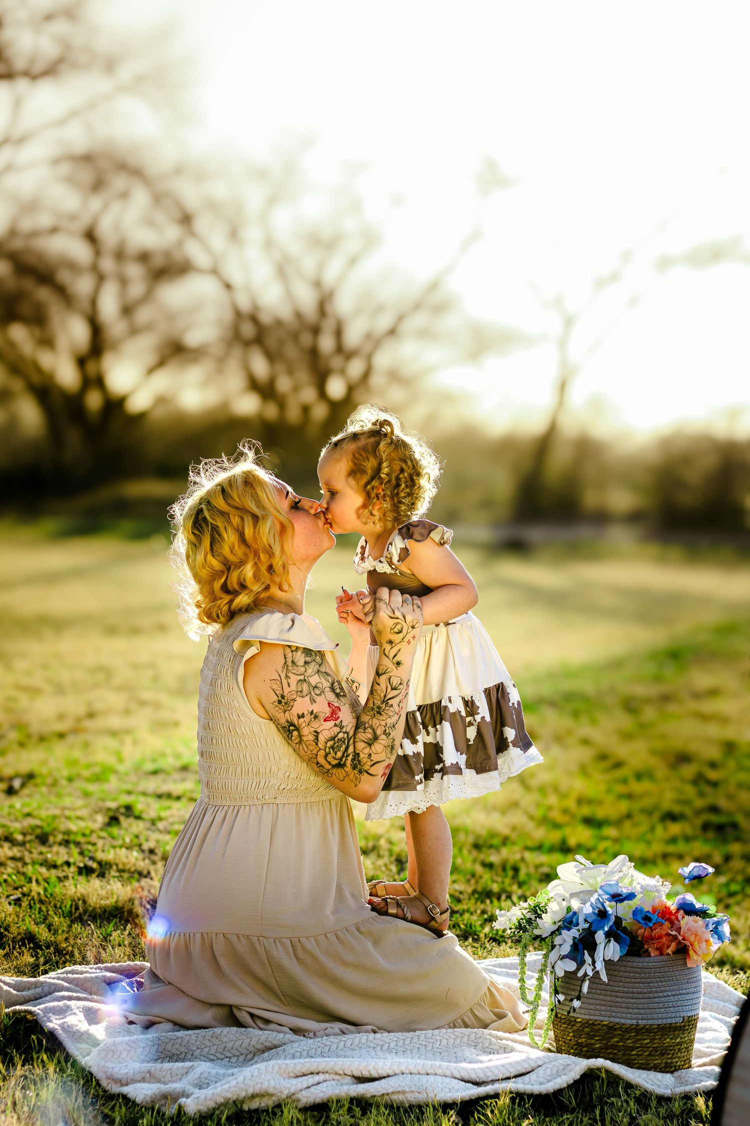 Tattooed mother and curly-haired toddler sharing a kiss in golden sunlit park.