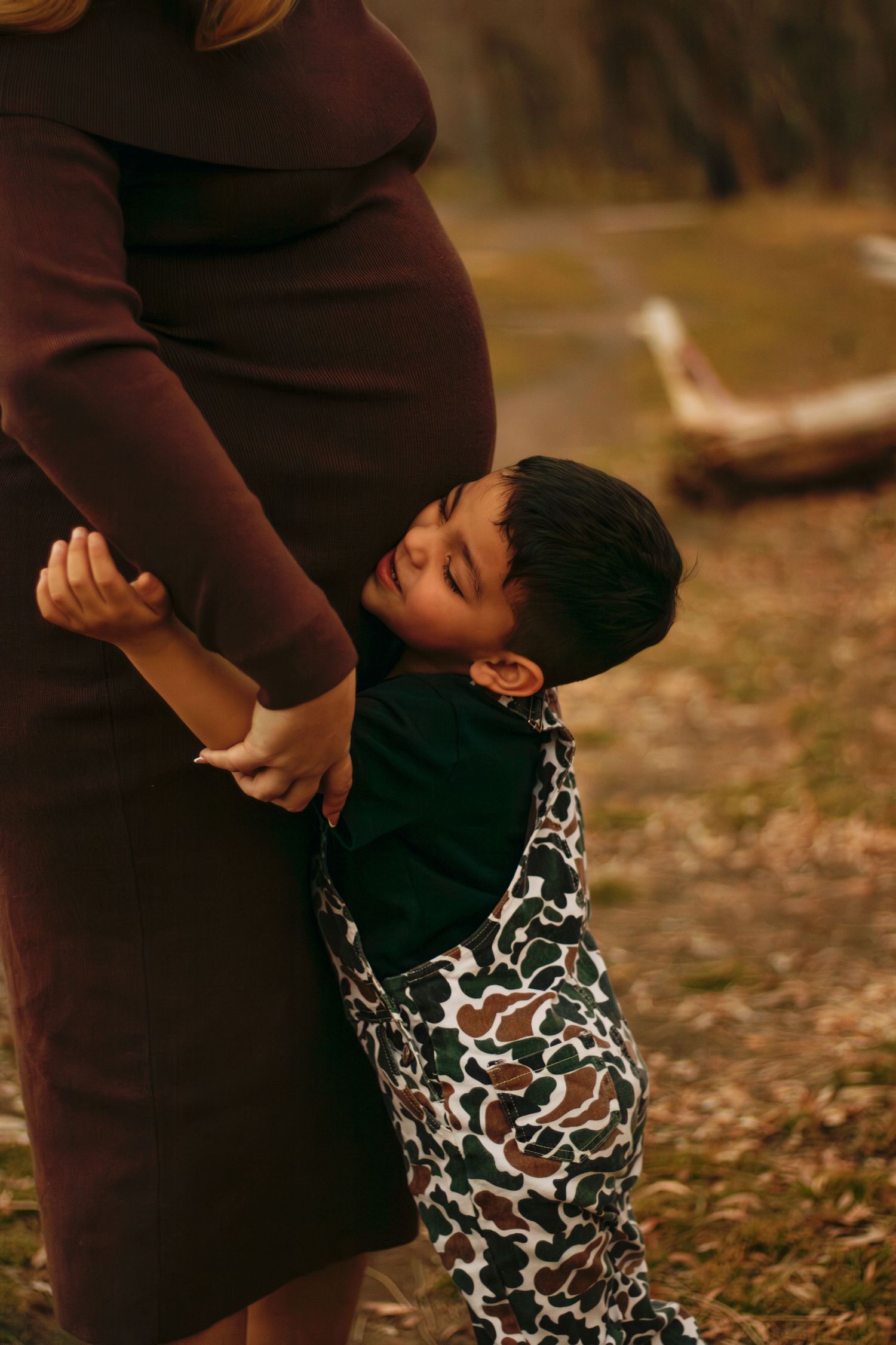 Young child lovingly hugging pregnant mothers baby bump outdoors in warm autumn setting.