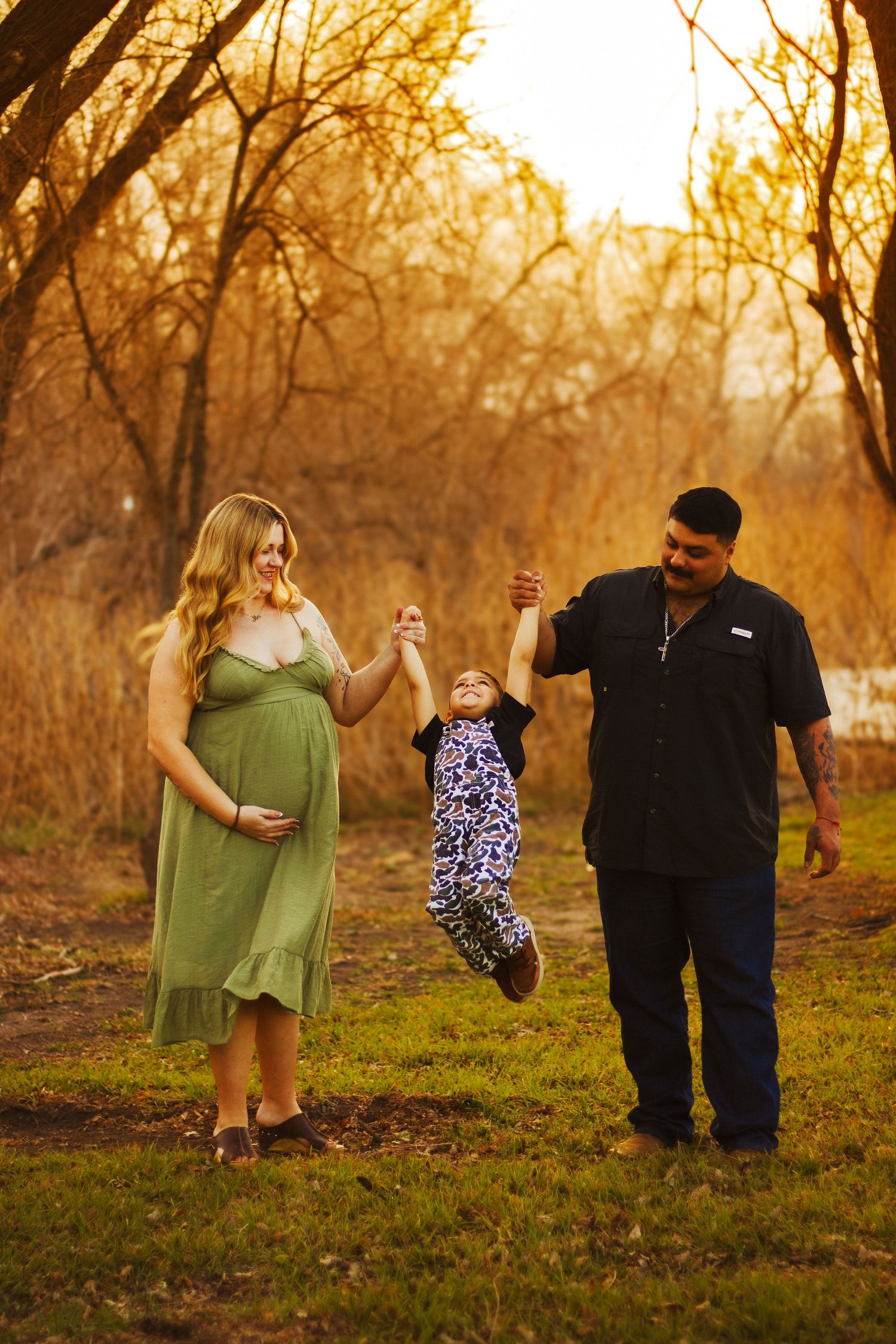 Joyful pregnant family swinging toddler between parents in golden hour autumn park.