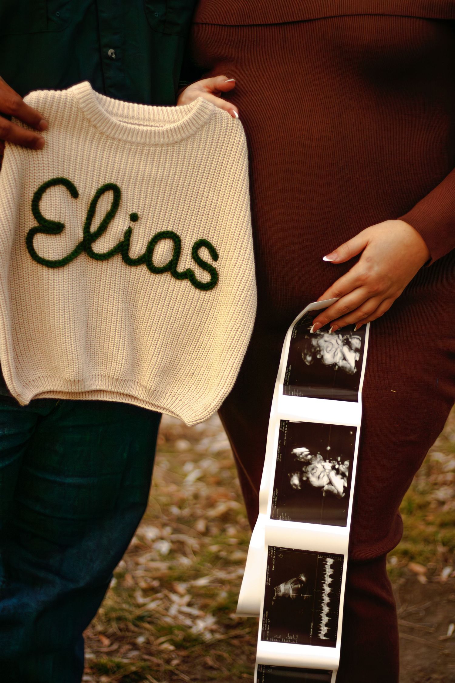 Close-up of baby name sweater reading Elias held beside pregnant belly with ultrasound strip.
