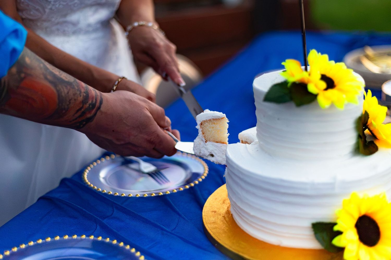 Couple cutting a white two-tier wedding cake decorated with sunflowers on a blue tablecloth.