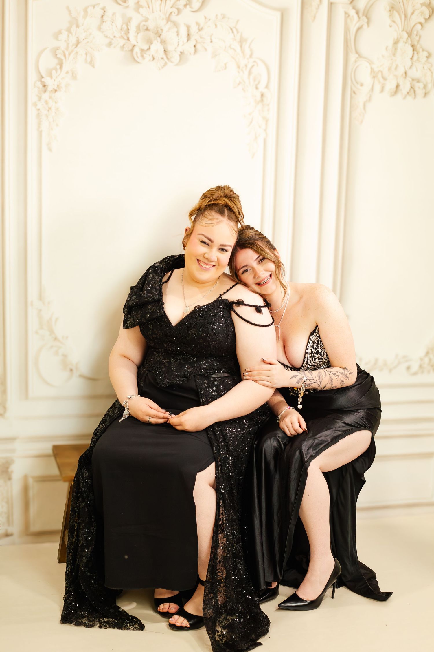 Two women in elegant black gowns sitting together, smiling, against an ornate white wall.