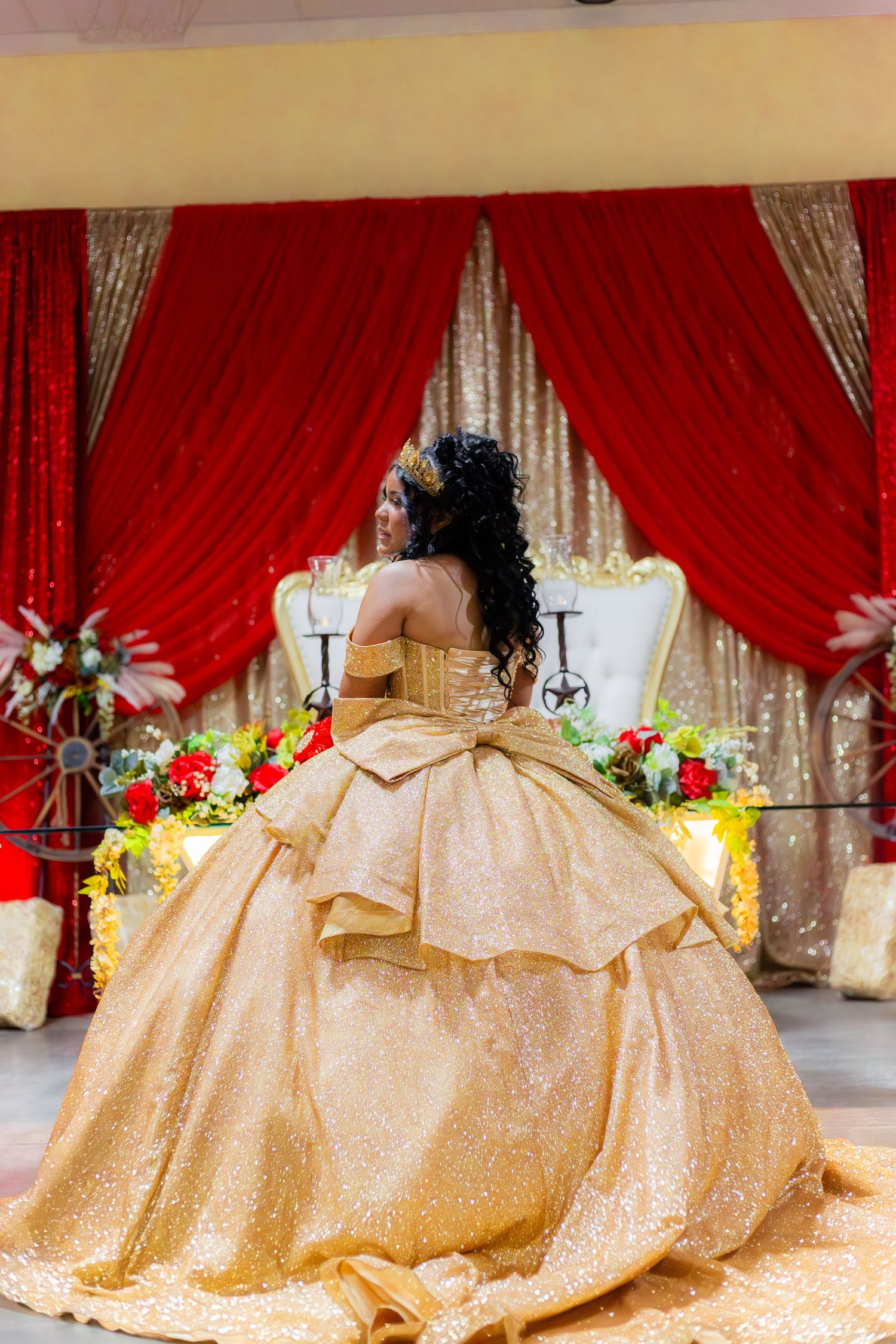 A young woman in a stunning gold ballgown poses at a quinceañera celebration with red curtains and floral decor.