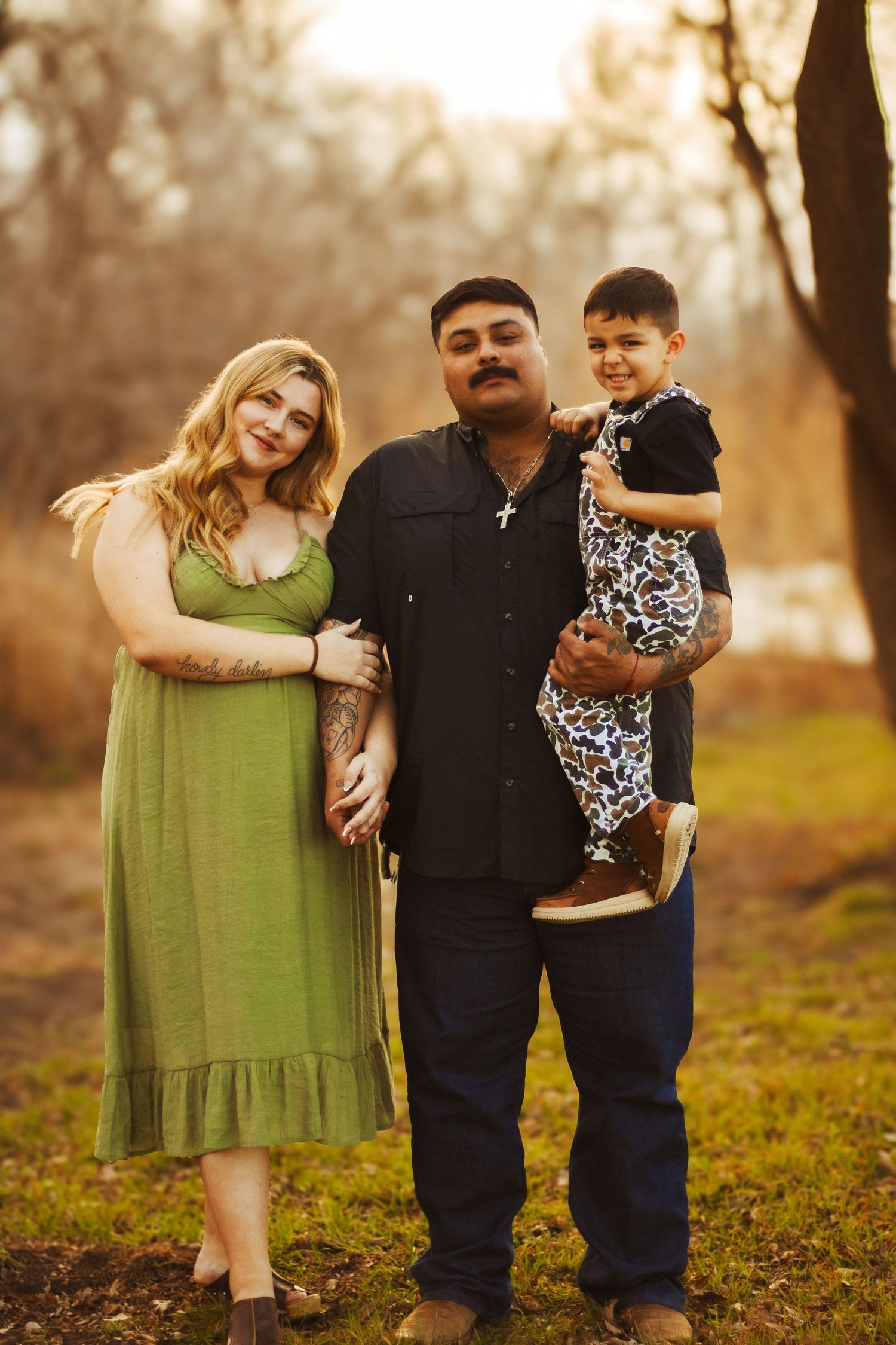 Happy interracial family posing outdoors at golden hour with man holding young child in nature.