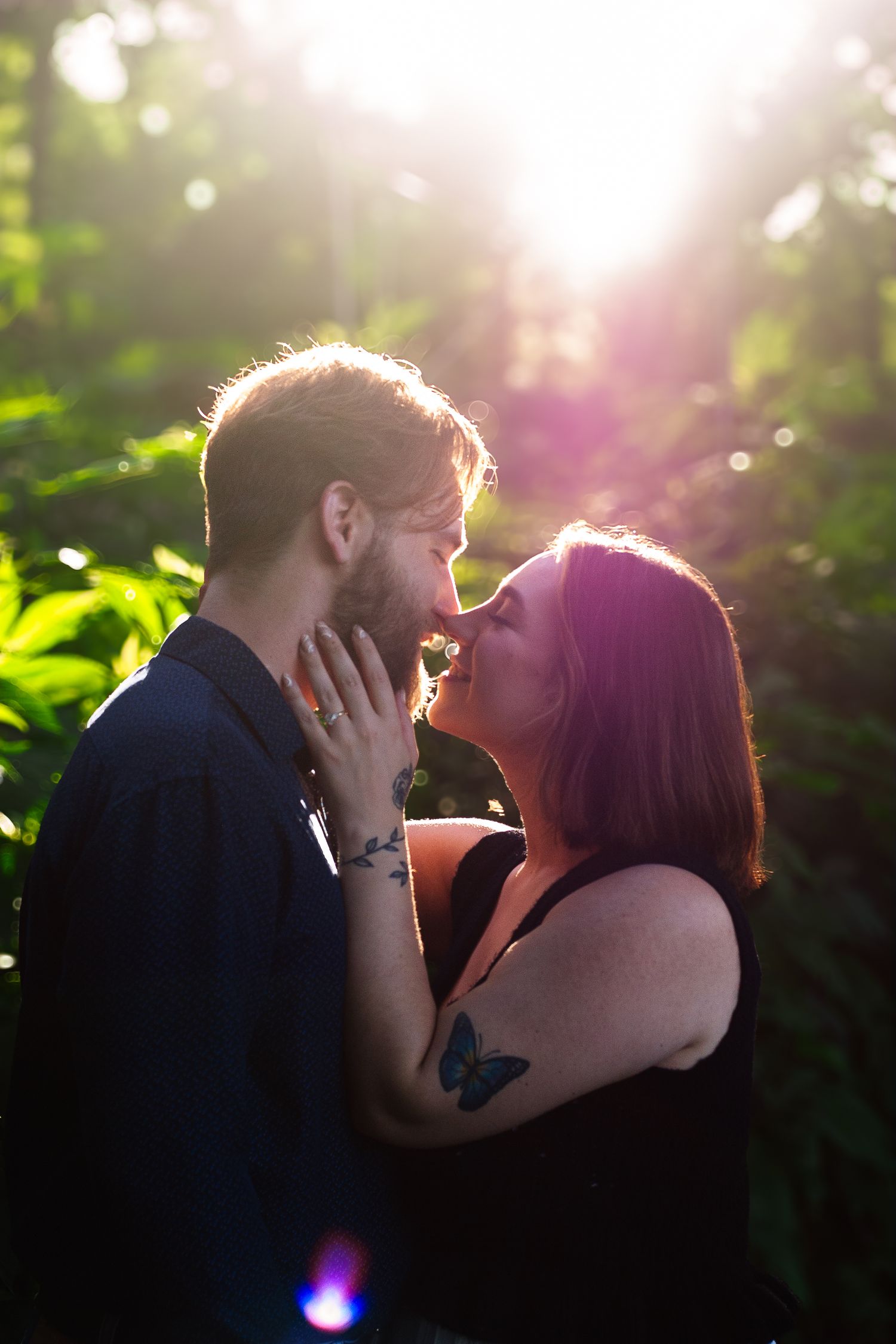 Couple sharing a romantic almost-kiss in sunlit forest with golden backlight and green foliage.
