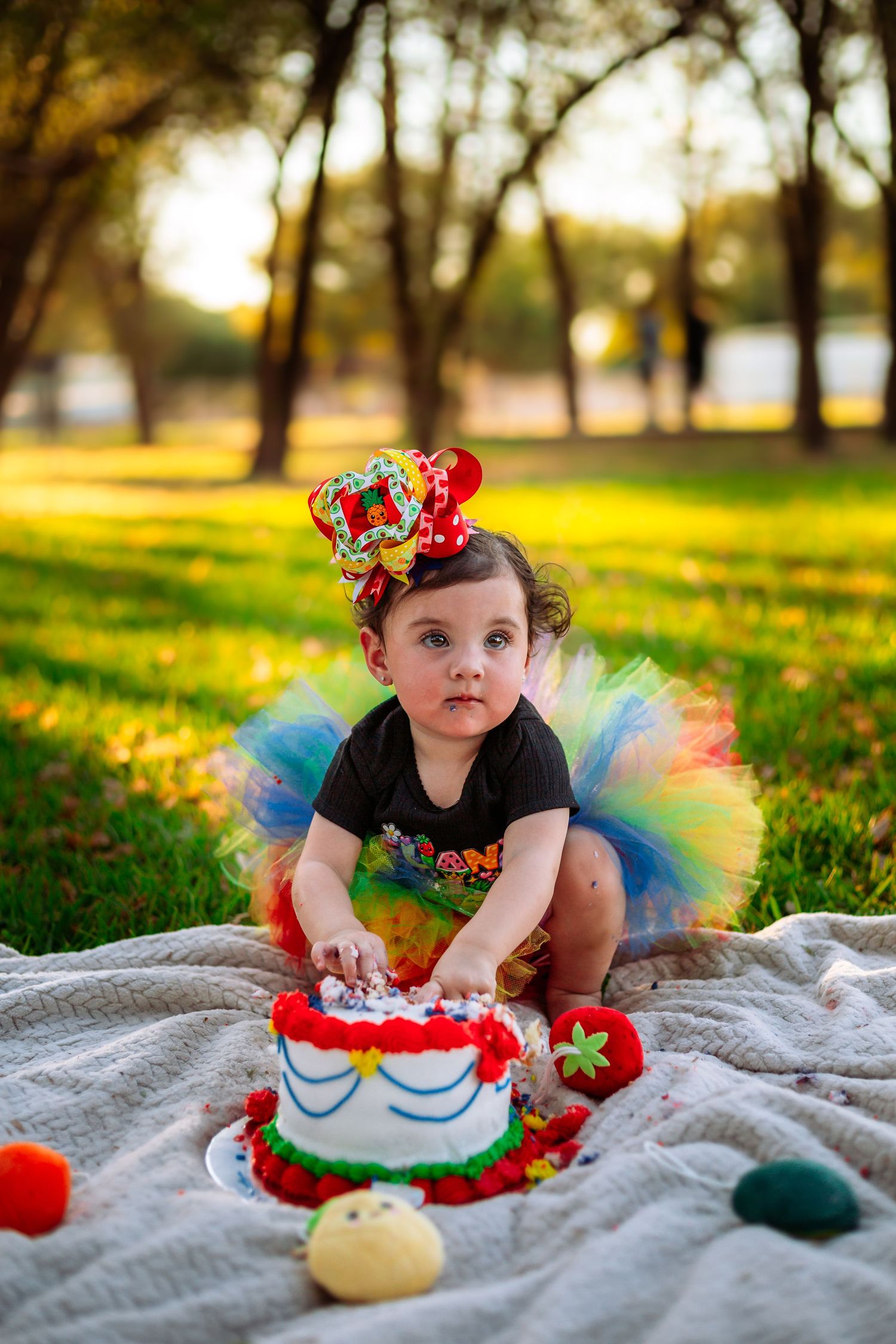 Toddler in colorful tutu and festive hat smashing first birthday cake outdoors on green grass in sunny park.