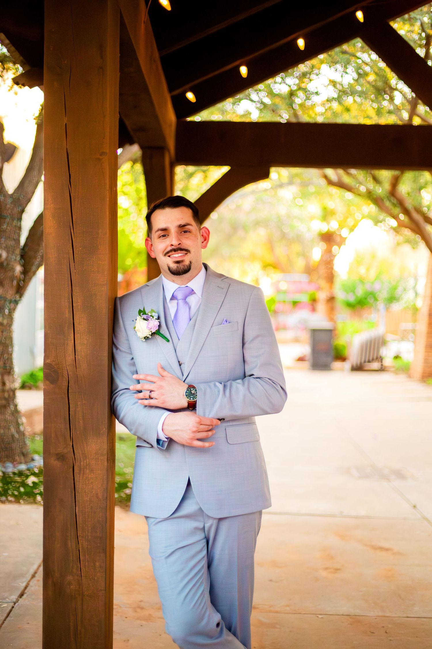 Groom in light gray suit with lavender tie and boutonniere leaning against wooden pergola post outdoors.