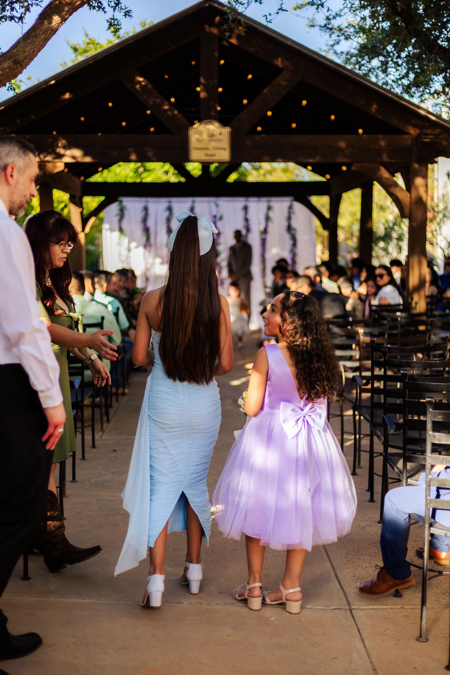 Woman in blue dress and girl in purple tutu walk down outdoor wedding aisle under string lights.