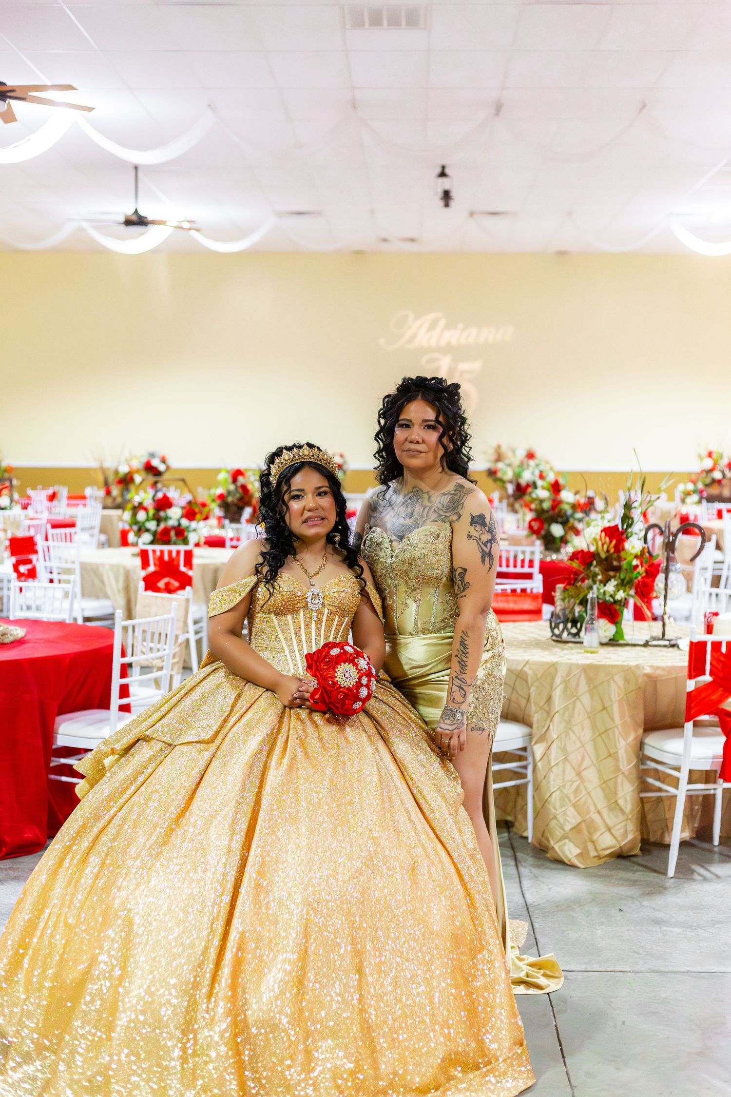Two women in gold gowns pose at a quinceañera celebration with red and gold decorated tables.
