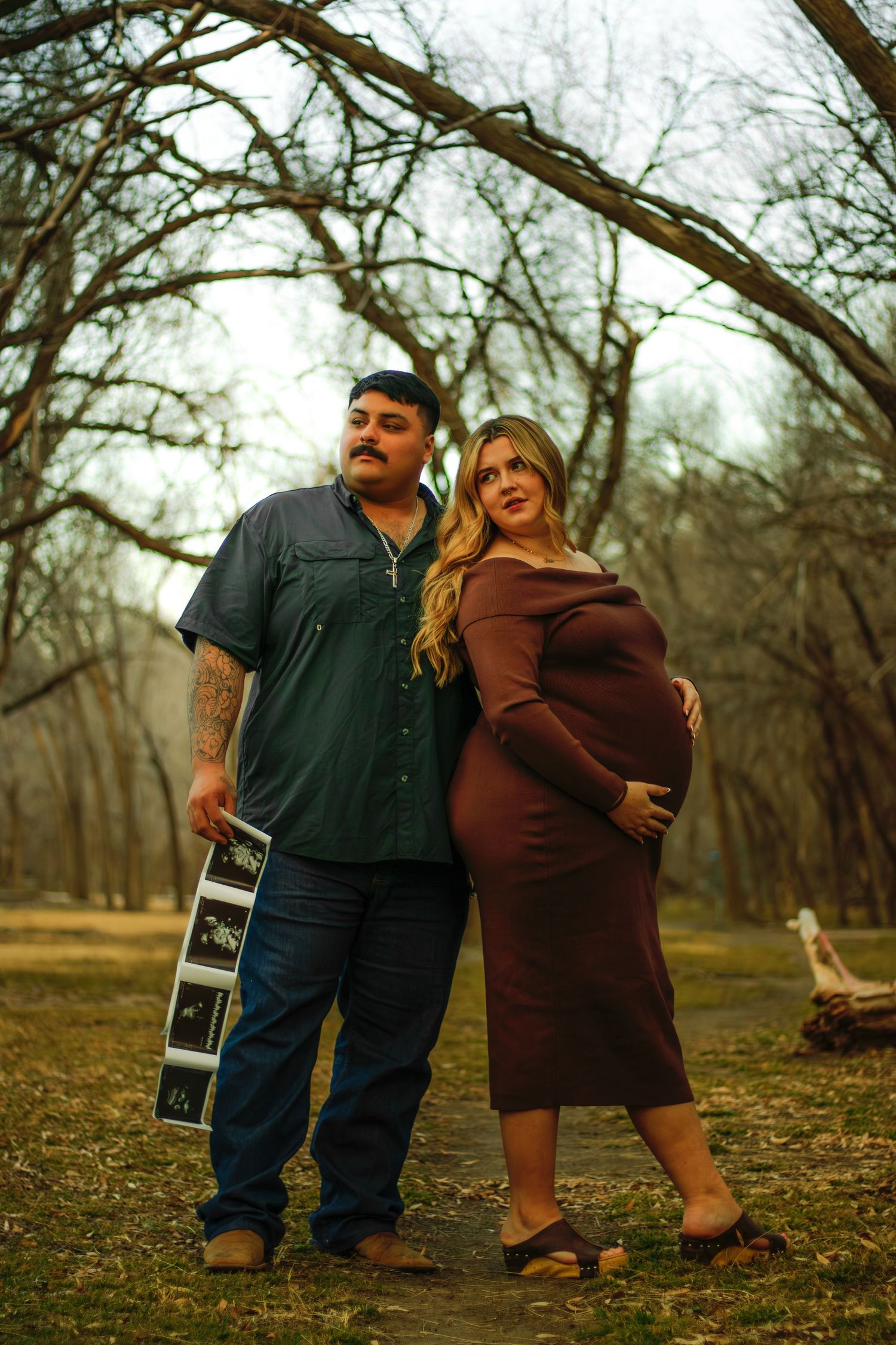 Expecting couple standing in bare winter park, holding ultrasound photos and cradling baby bump.