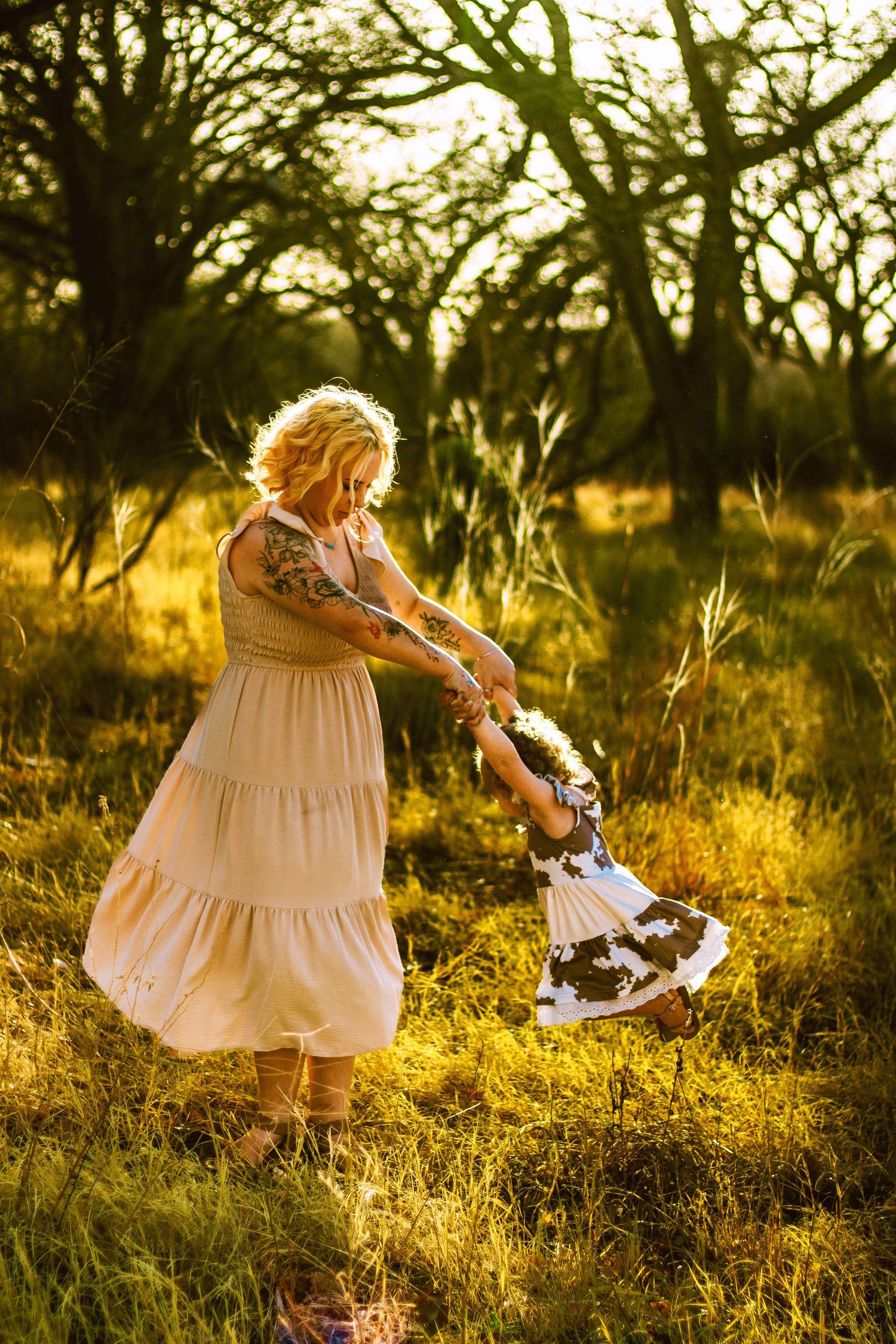 Tattooed blonde woman in white dress twirling young child in cow-print outfit in golden sunlit field.