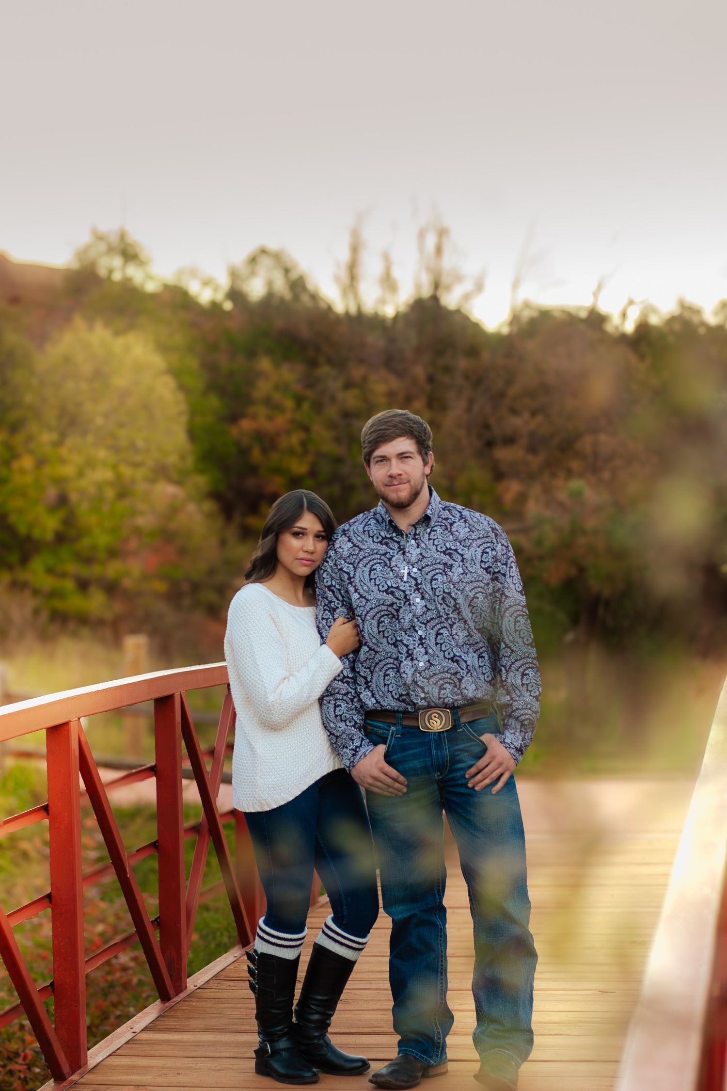 Young couple posing together on a rustic red bridge surrounded by autumn foliage at golden hour.