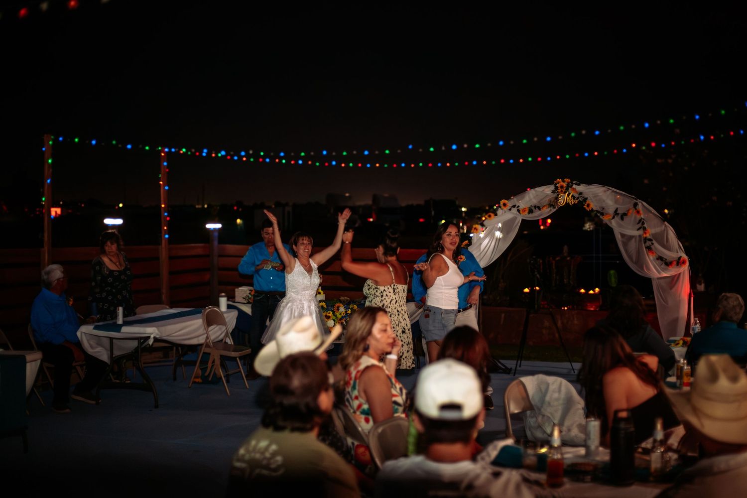 Wedding couple celebrates under a floral arch at an outdoor nighttime reception with string lights.
