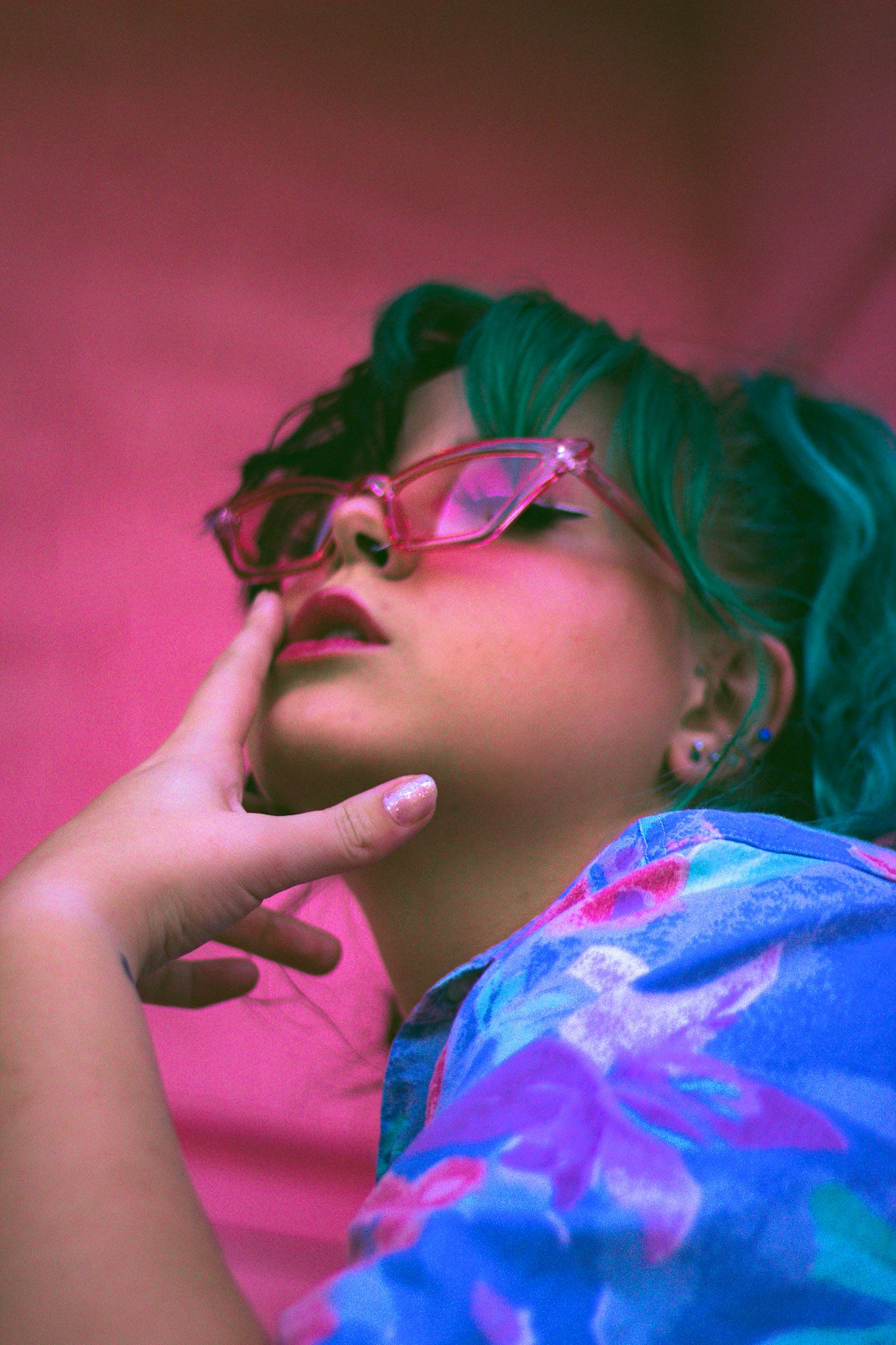 Close-up of woman with green hair and pink cat-eye glasses against a vibrant pink background.