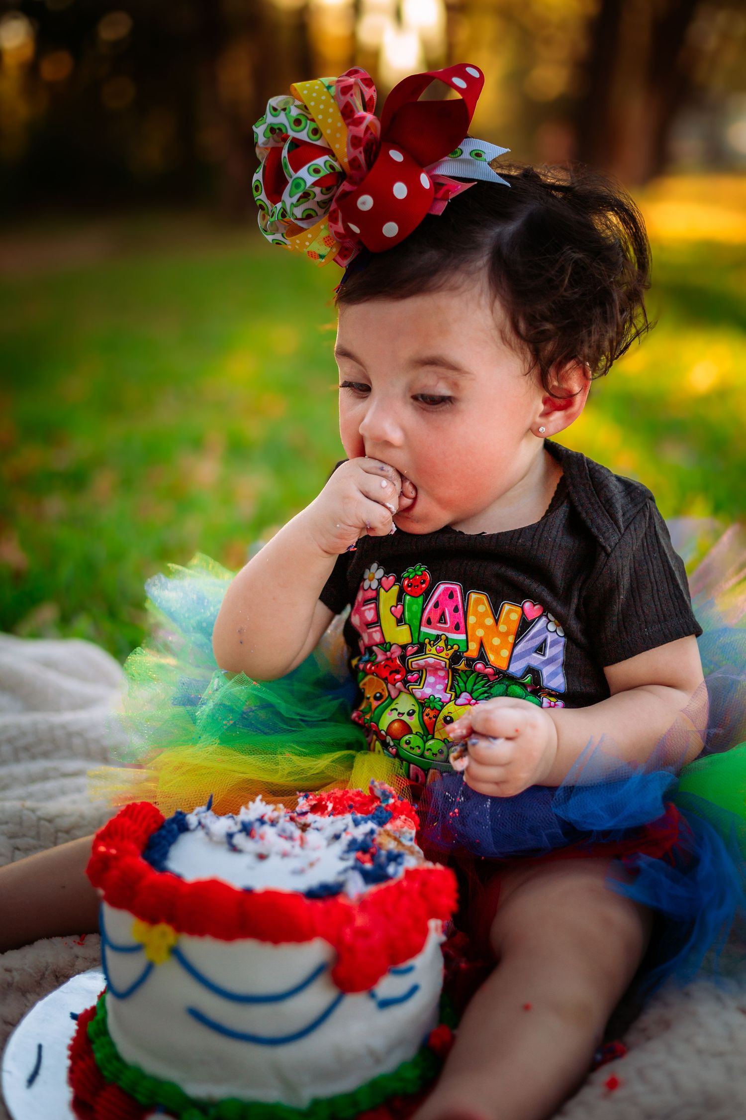 Baby girl in colorful tutu and bow headband eating her first birthday cake outdoors in a sunny park.