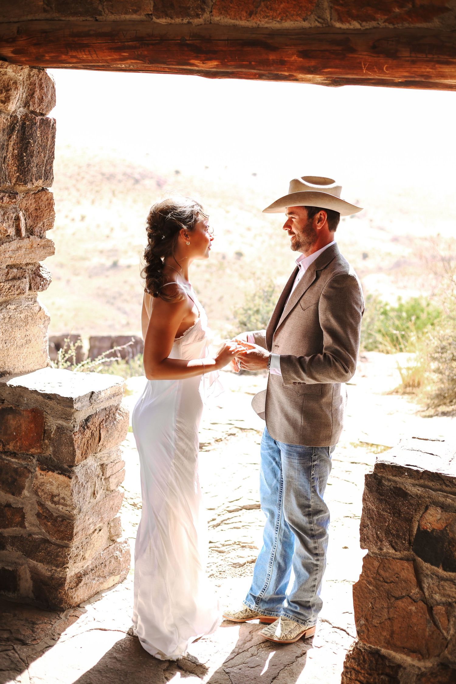 Bride and groom exchange vows in rustic stone chapel with desert mountain backdrop, groom wearing cowboy hat and jeans.