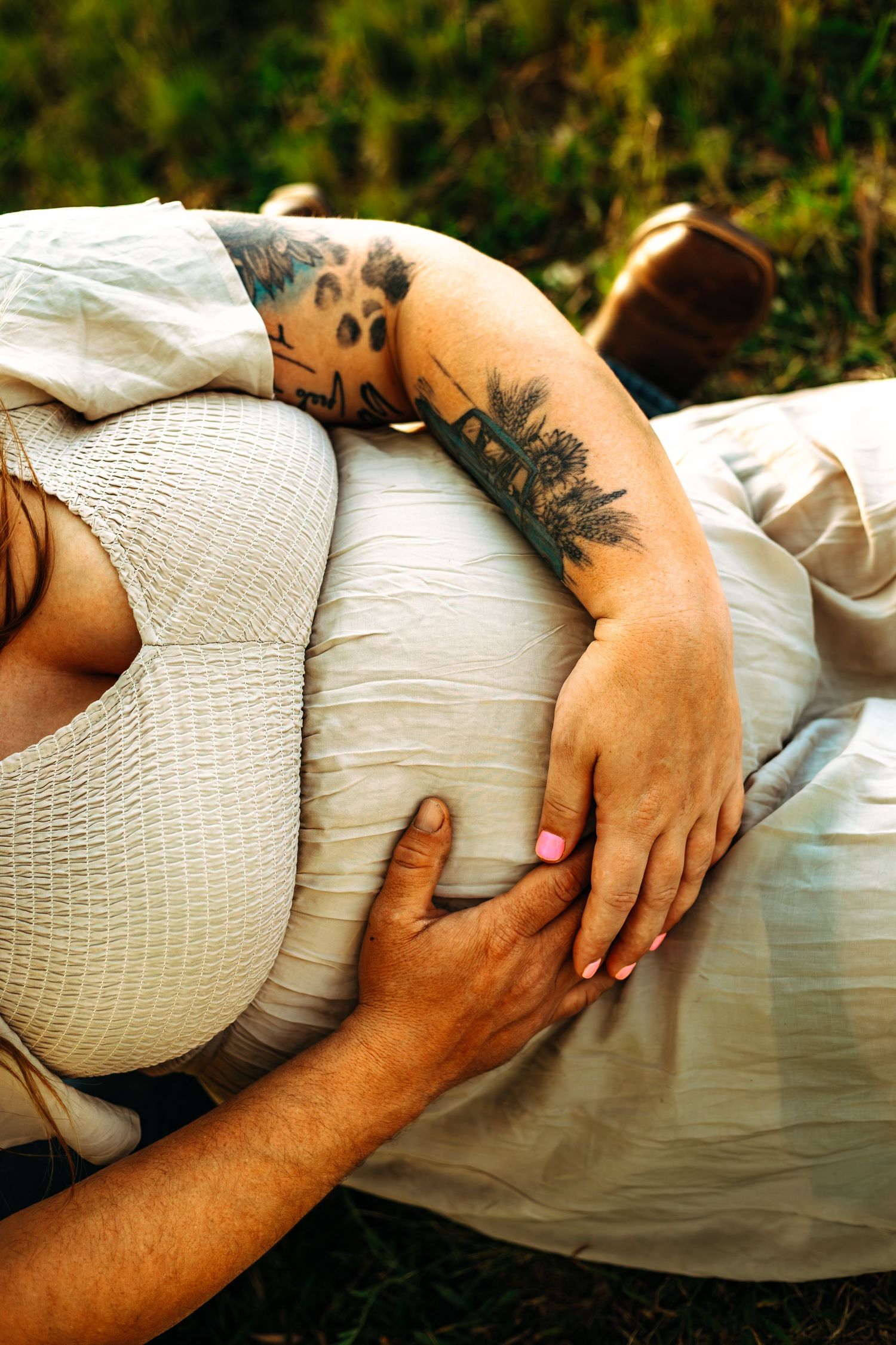 Pregnant woman in white dress with tattoos cradled by partner outdoors in nature.