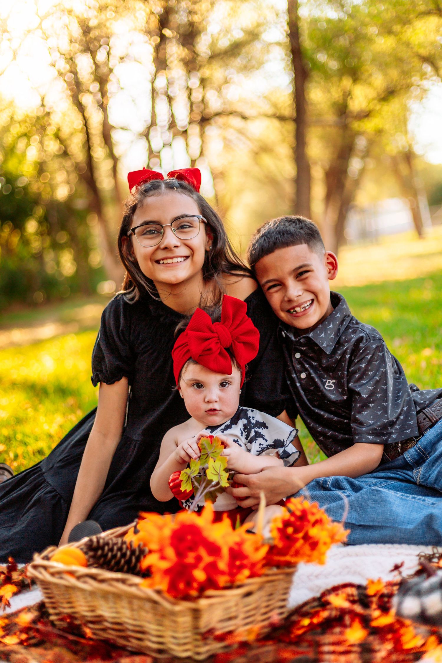 Three siblings pose for a fall family photo outdoors, with two older kids smiling beside a baby holding autumn leaves.