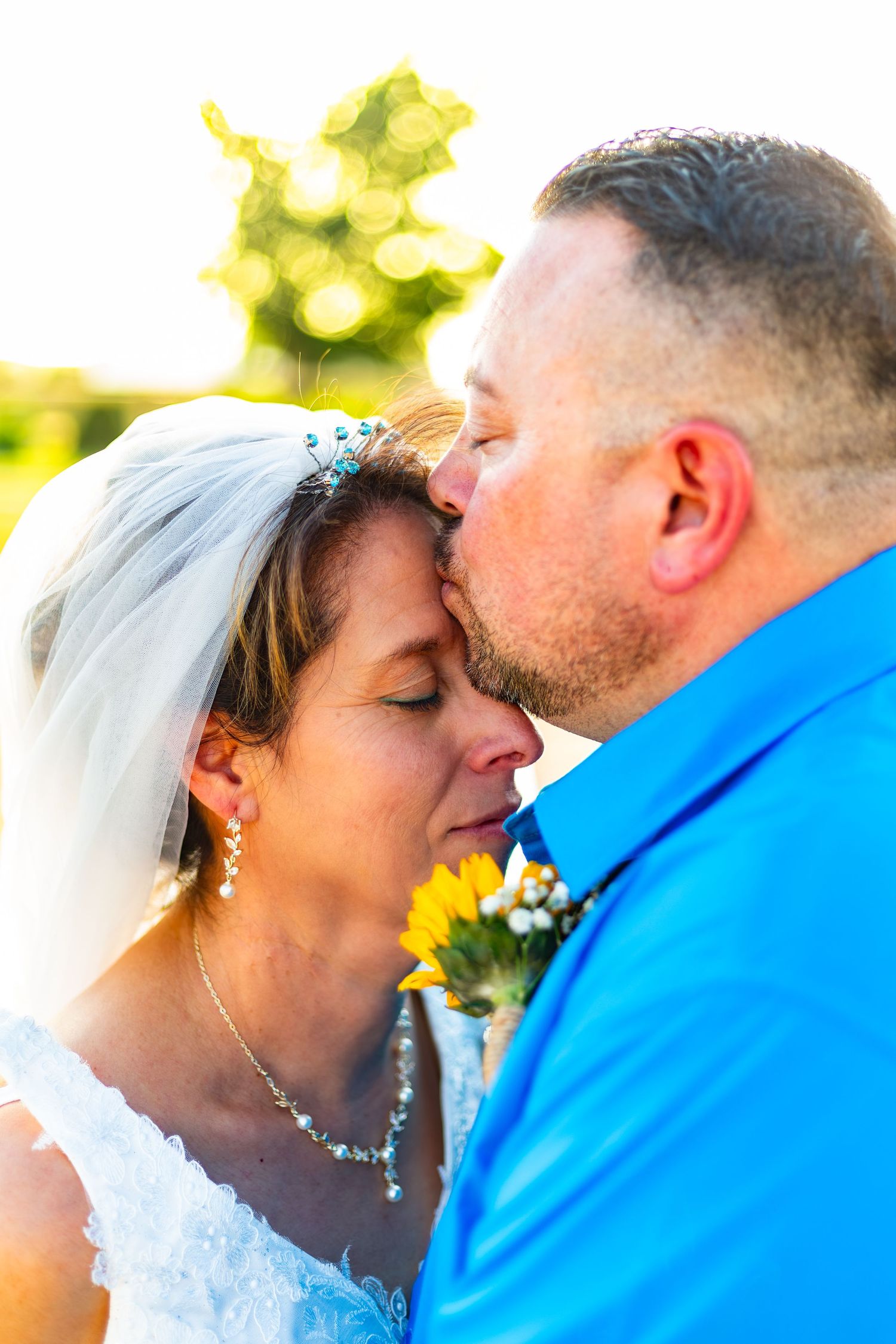 Groom in blue shirt tenderly kissing bride wearing veil and sunflower bouquet in golden outdoor wedding photo.