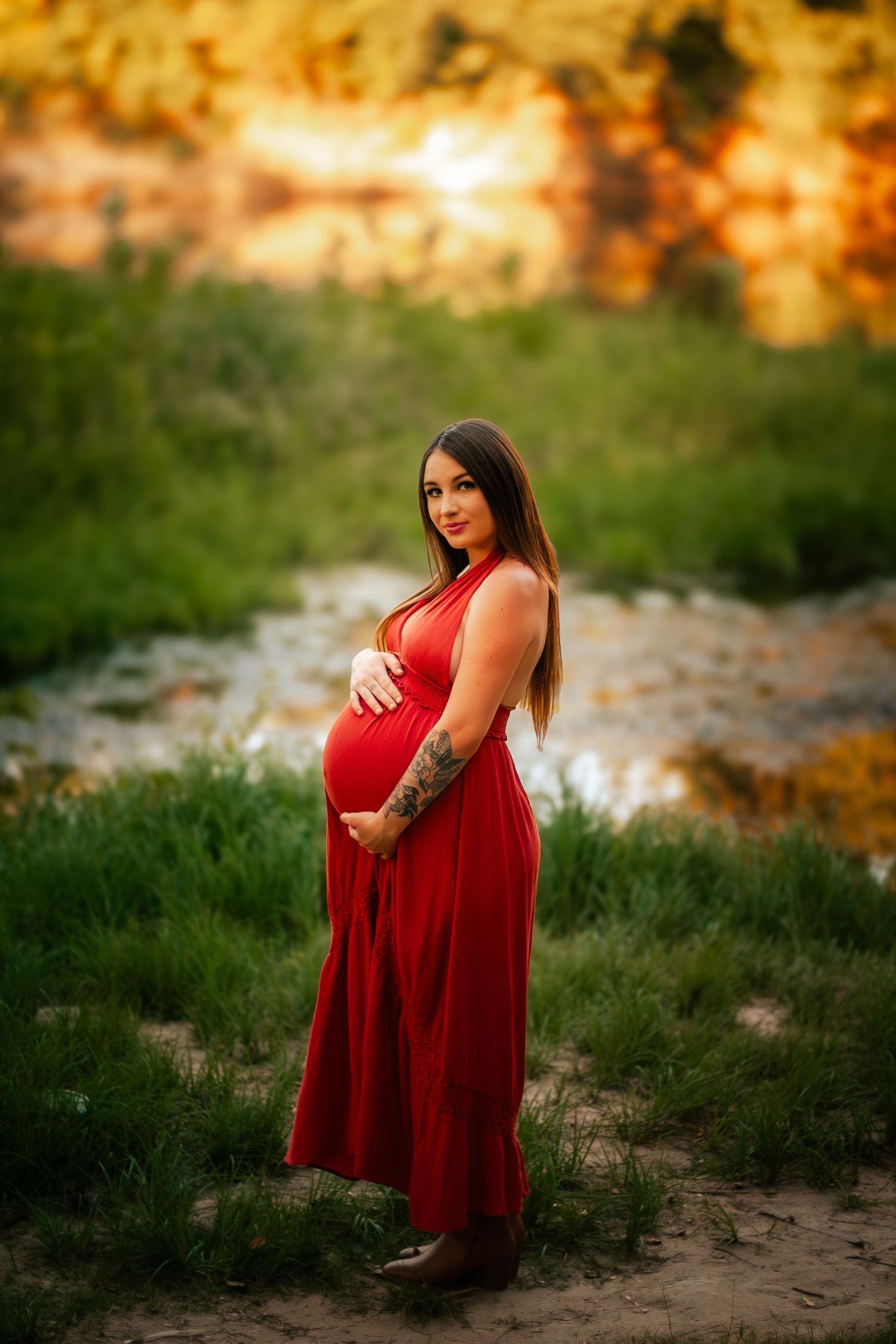 Beautiful pregnant woman in flowing red dress standing in sunlit green field at sunset.