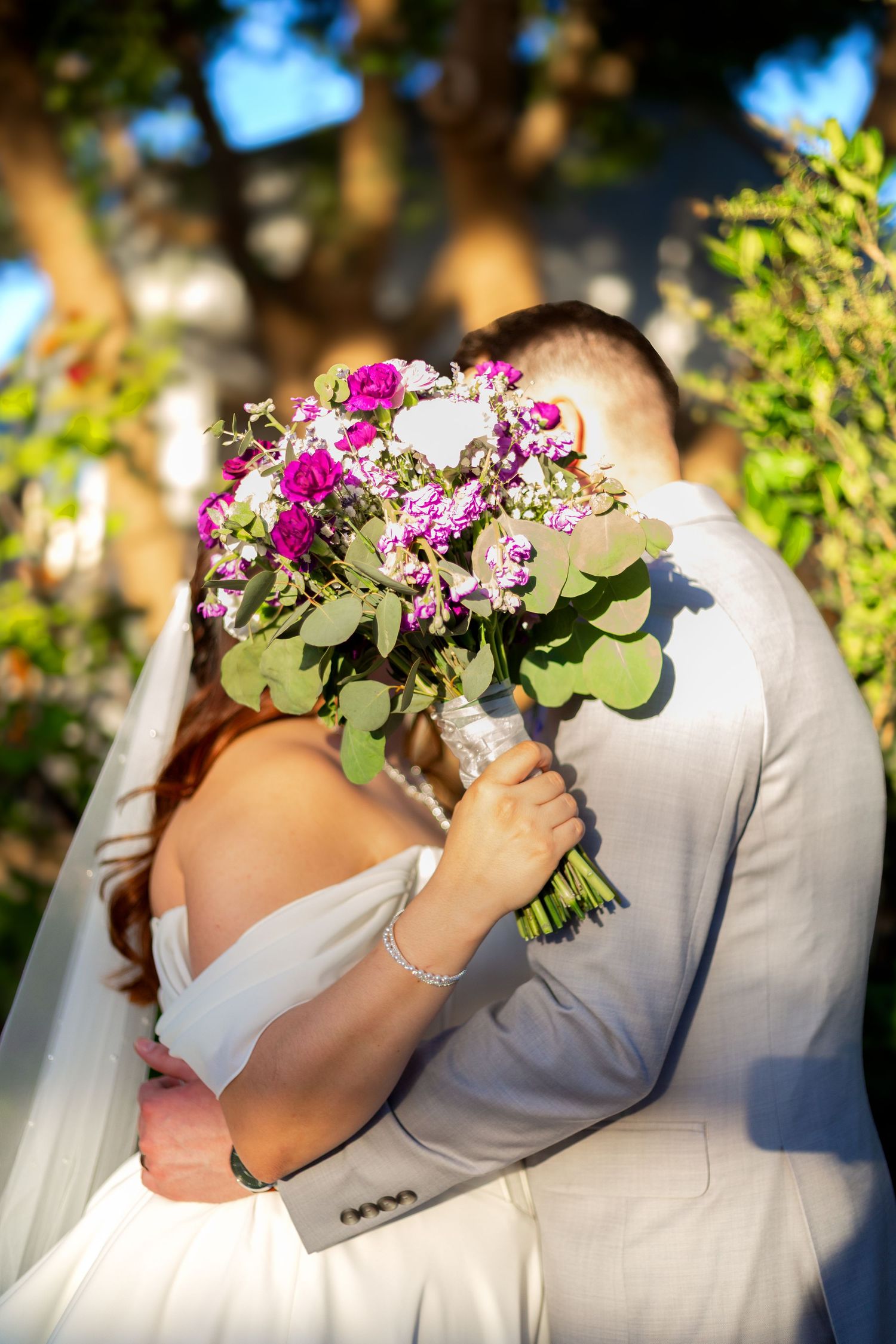 Bride and groom kissing behind a vibrant pink and purple wedding bouquet in an outdoor garden setting.