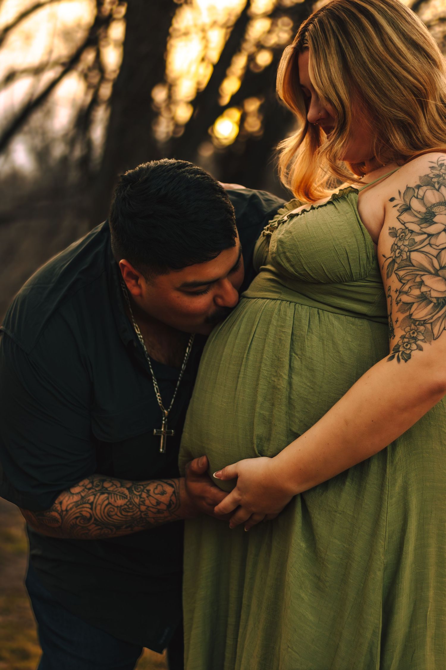 Man kissing pregnant woman's belly in golden sunset forest setting.