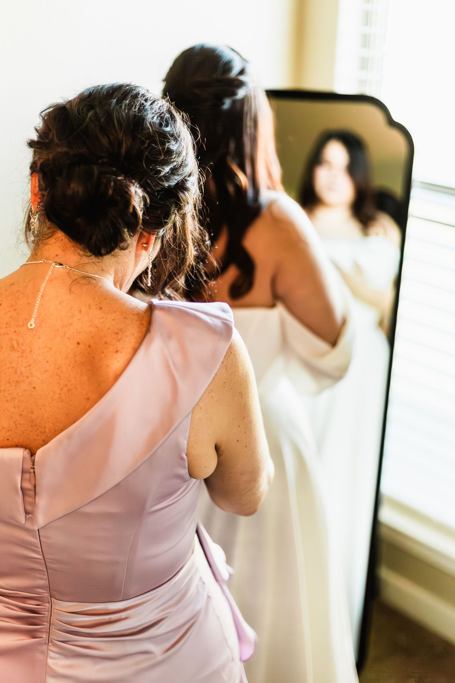 Bridesmaid in pink dress helps bride in white gown prepare, reflected in elegant mirror near window.
