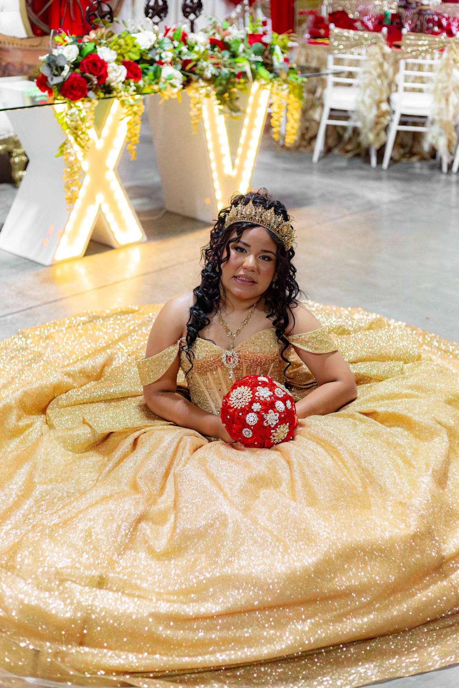 Young girl in gold quinceañera gown with crown holds red bouquet, seated on dance floor at XV celebration.