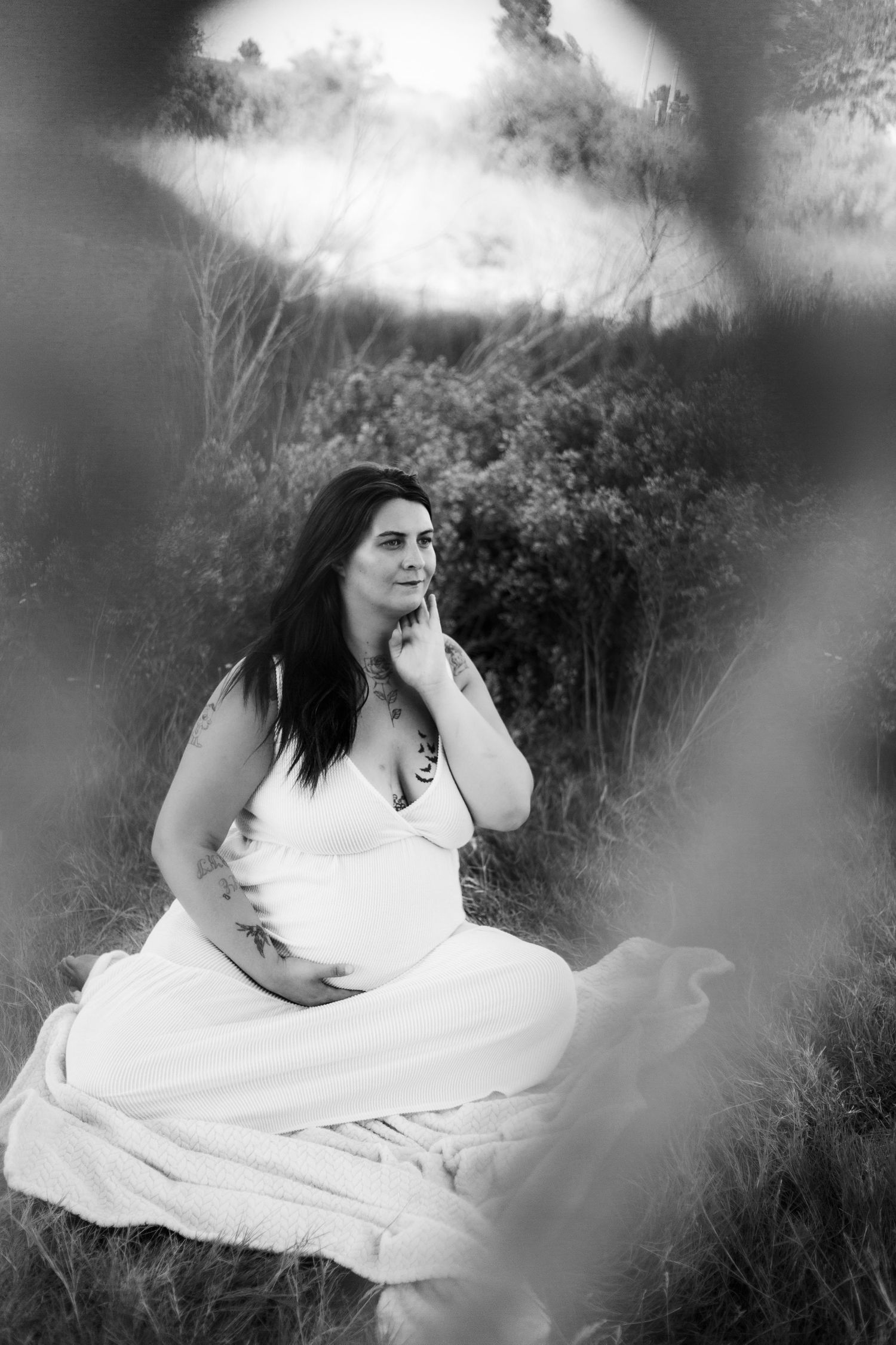 Black and white photo of a pregnant woman in a white dress sitting in a field, touching her face thoughtfully.