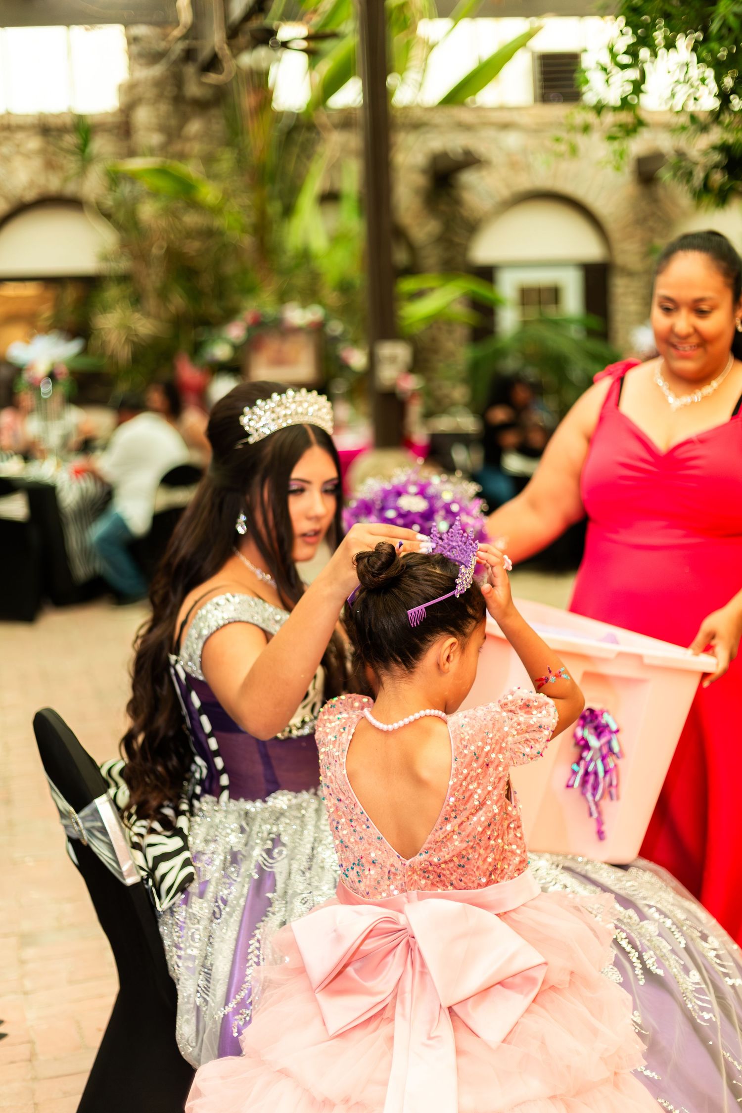 A quinceañera girl places a tiara on a young girl in a pink sequin dress at an outdoor celebration.