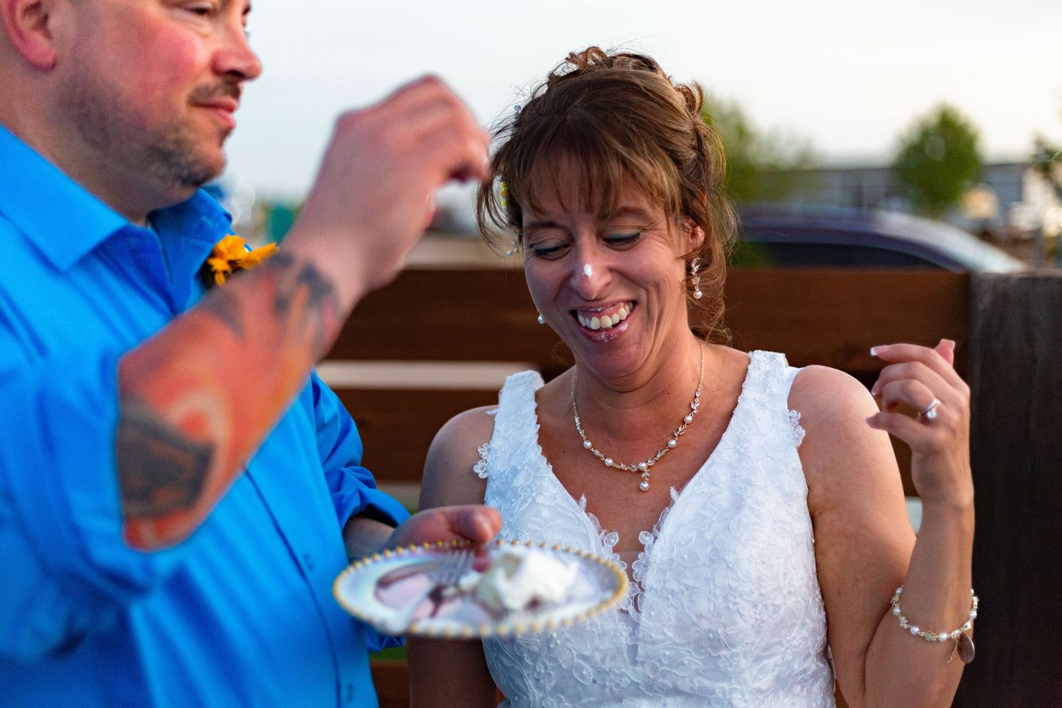 Bride in white lace dress laughs joyfully as groom in blue shirt with tattoos feeds her wedding cake outdoors.