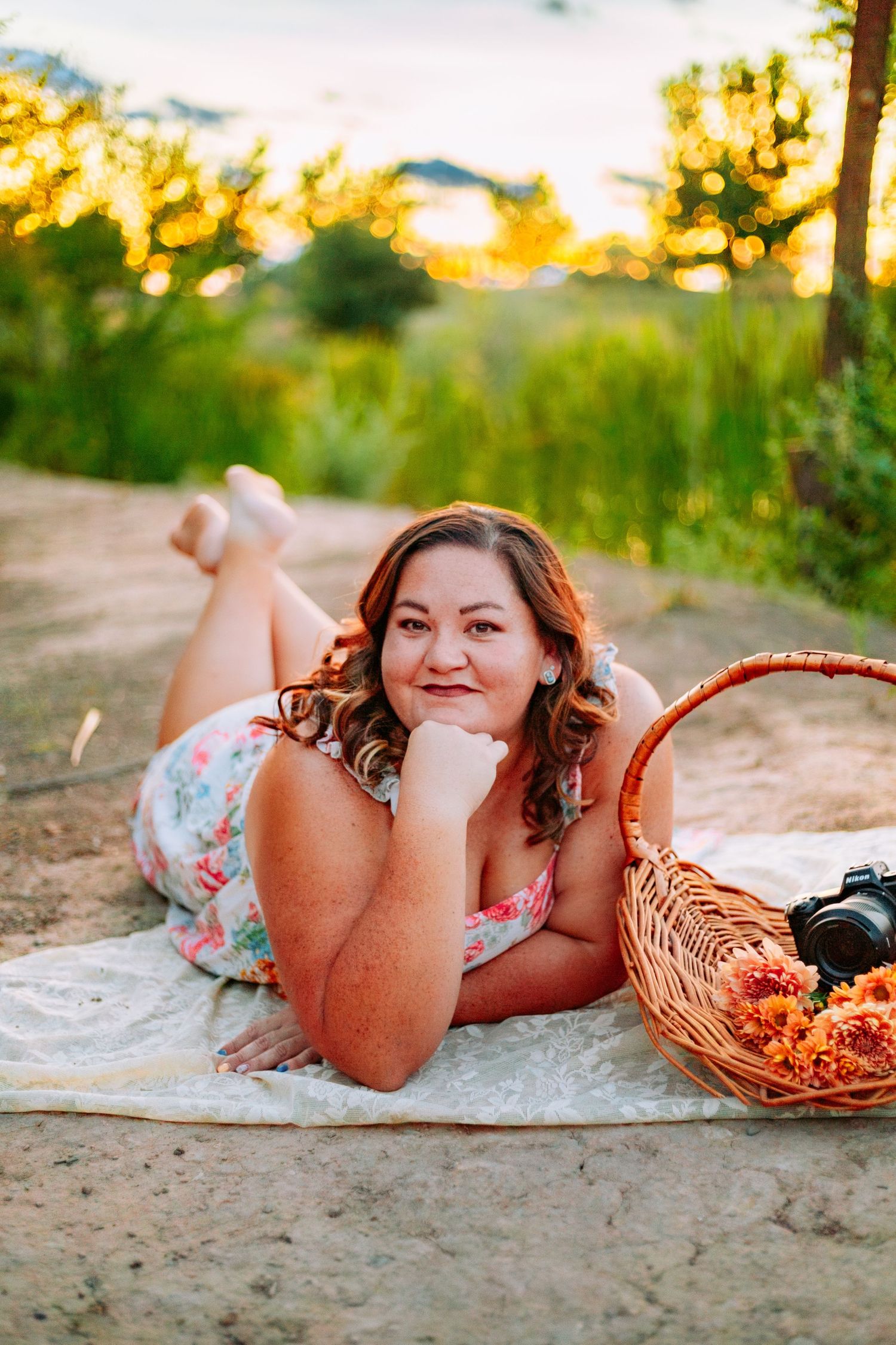 Woman in floral dress lying on blanket outdoors at sunset, smiling beside a wicker basket with camera.