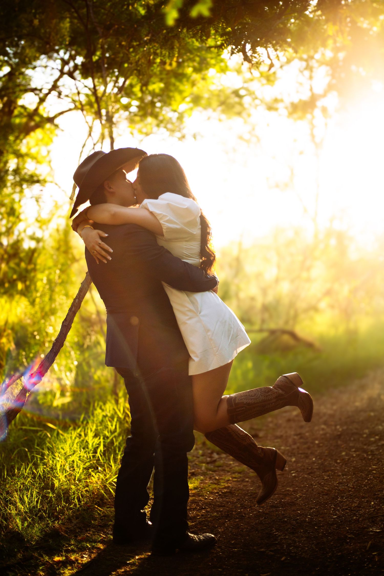 A couple shares a romantic kiss at sunset in a lush green forest, the woman lifting her leg in a white dress and cowboy boots.