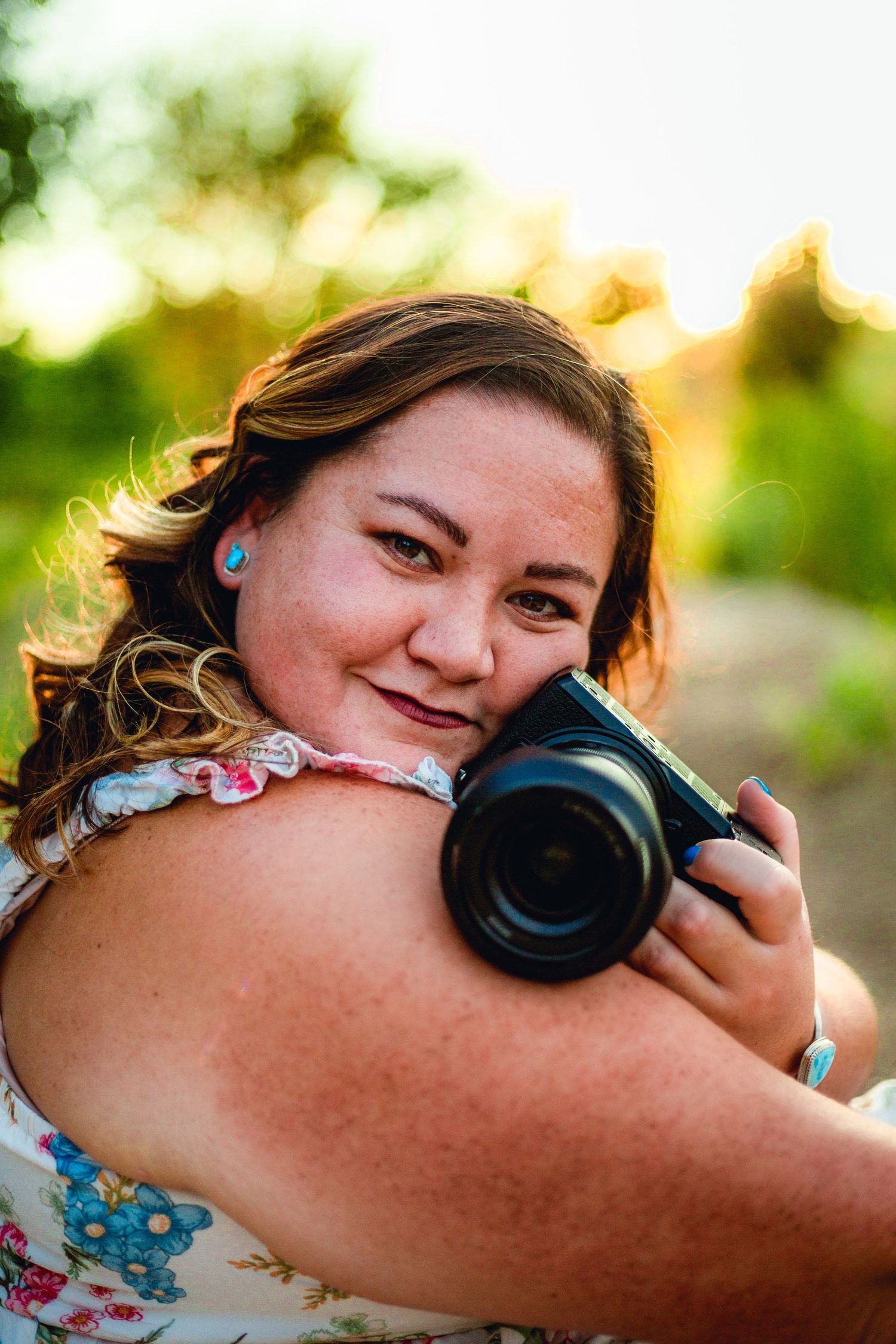 Young woman with freckles smiles while holding a camera outdoors in golden hour sunlight.