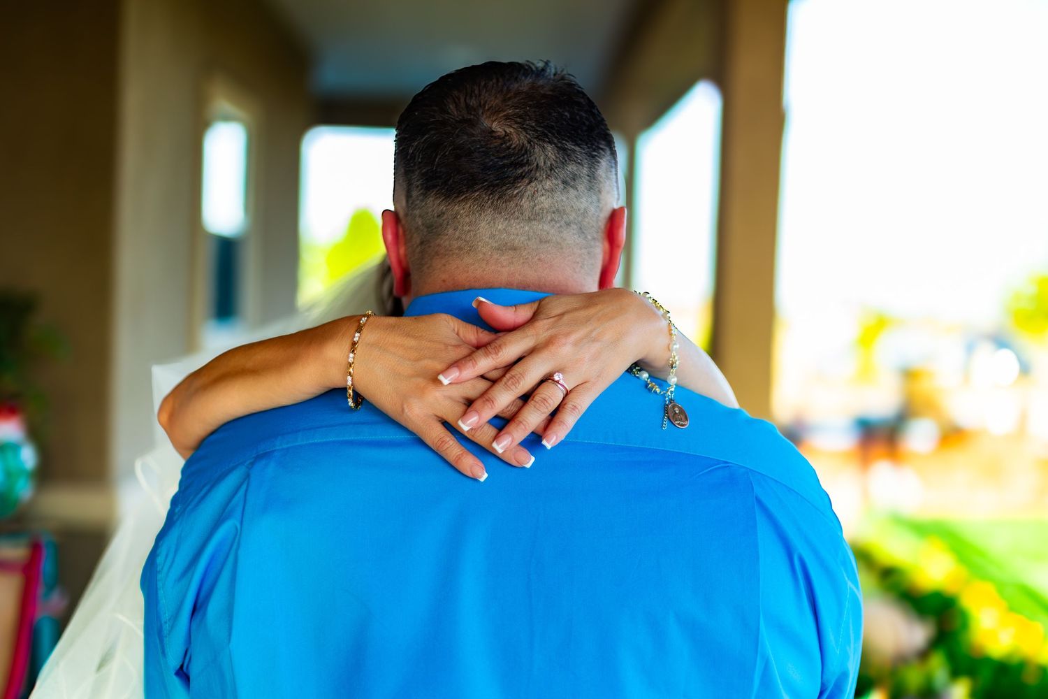 Couple embracing in a warm hug outdoors, woman's hand with gold bracelet resting on man's neck in blue shirt.