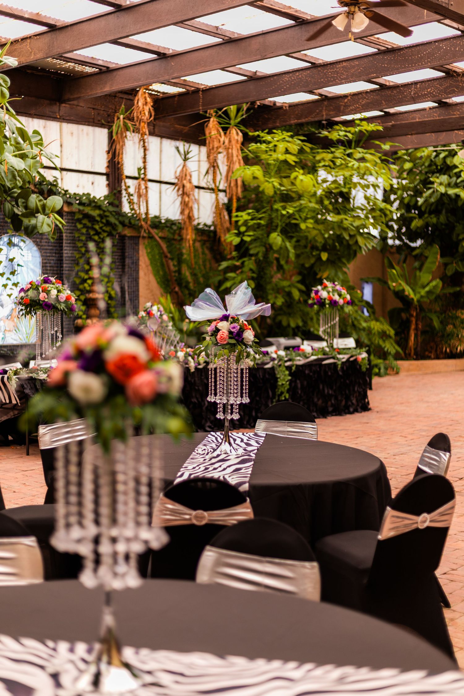 Elegant event venue with black tablecloths, crystal centerpieces, floral arrangements, and lush greenery under a pergola.