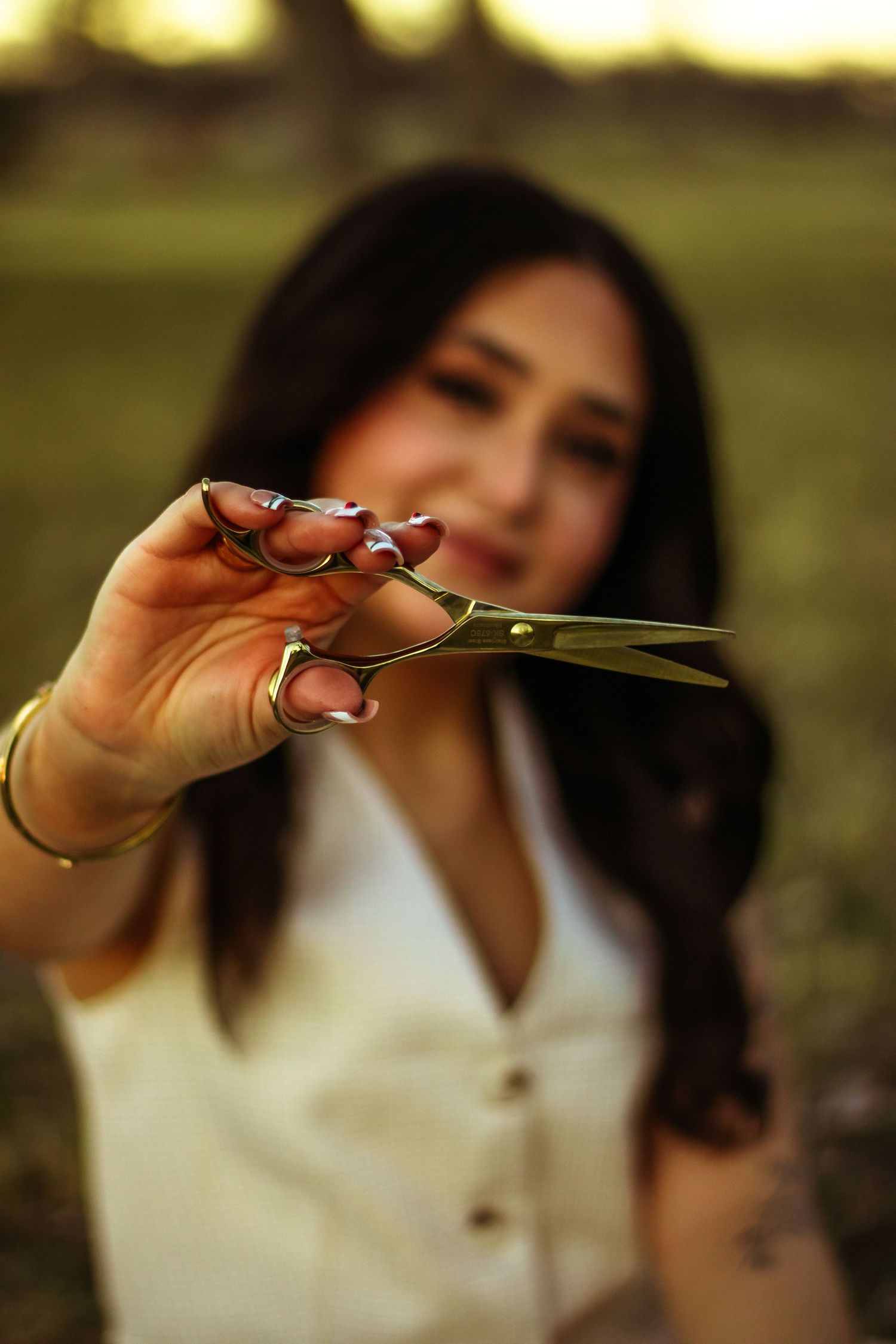 A woman with dark hair holds gold scissors toward the camera outdoors, wearing a white top with red manicured nails.