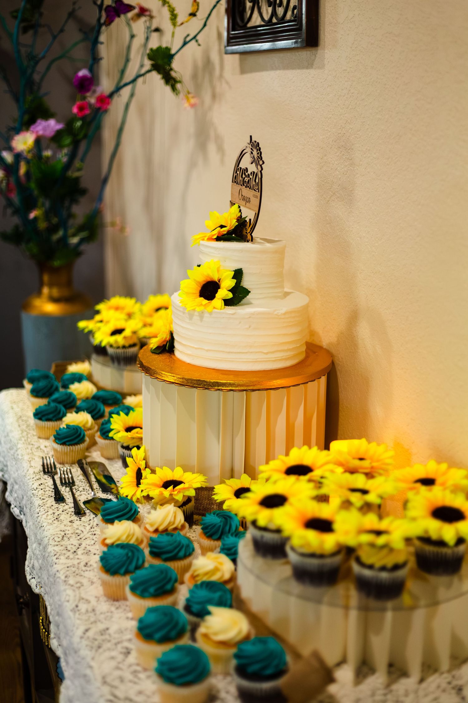 Elegant two-tier white wedding cake with sunflowers surrounded by teal and white cupcakes.