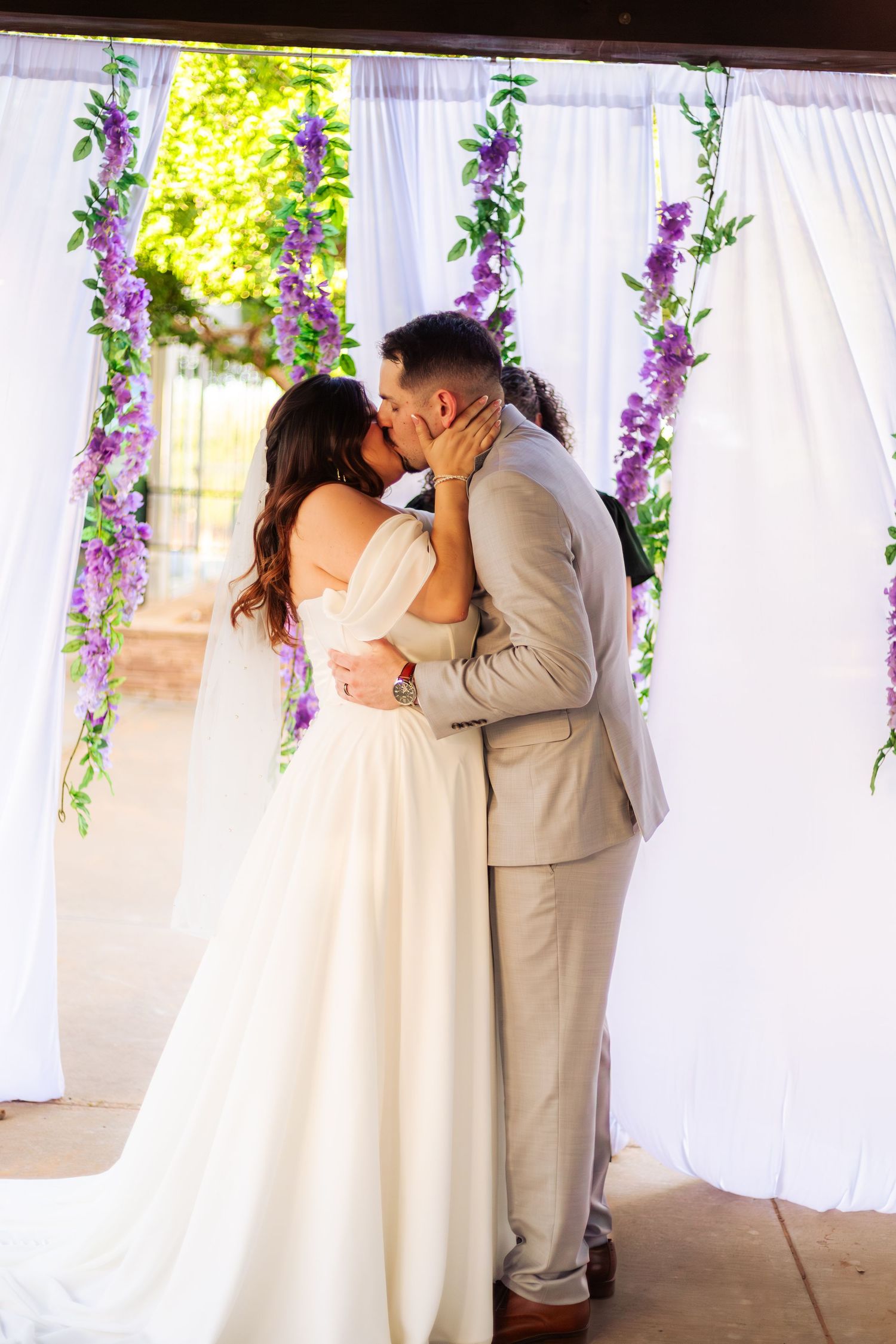 Bride and groom share first kiss at wedding altar adorned with white drapes and purple floral garlands.