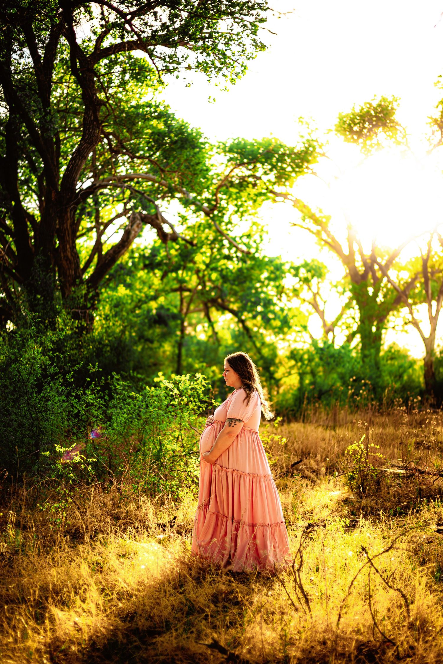 Pregnant woman in flowing pink dress standing in sunlit forest at sunset.