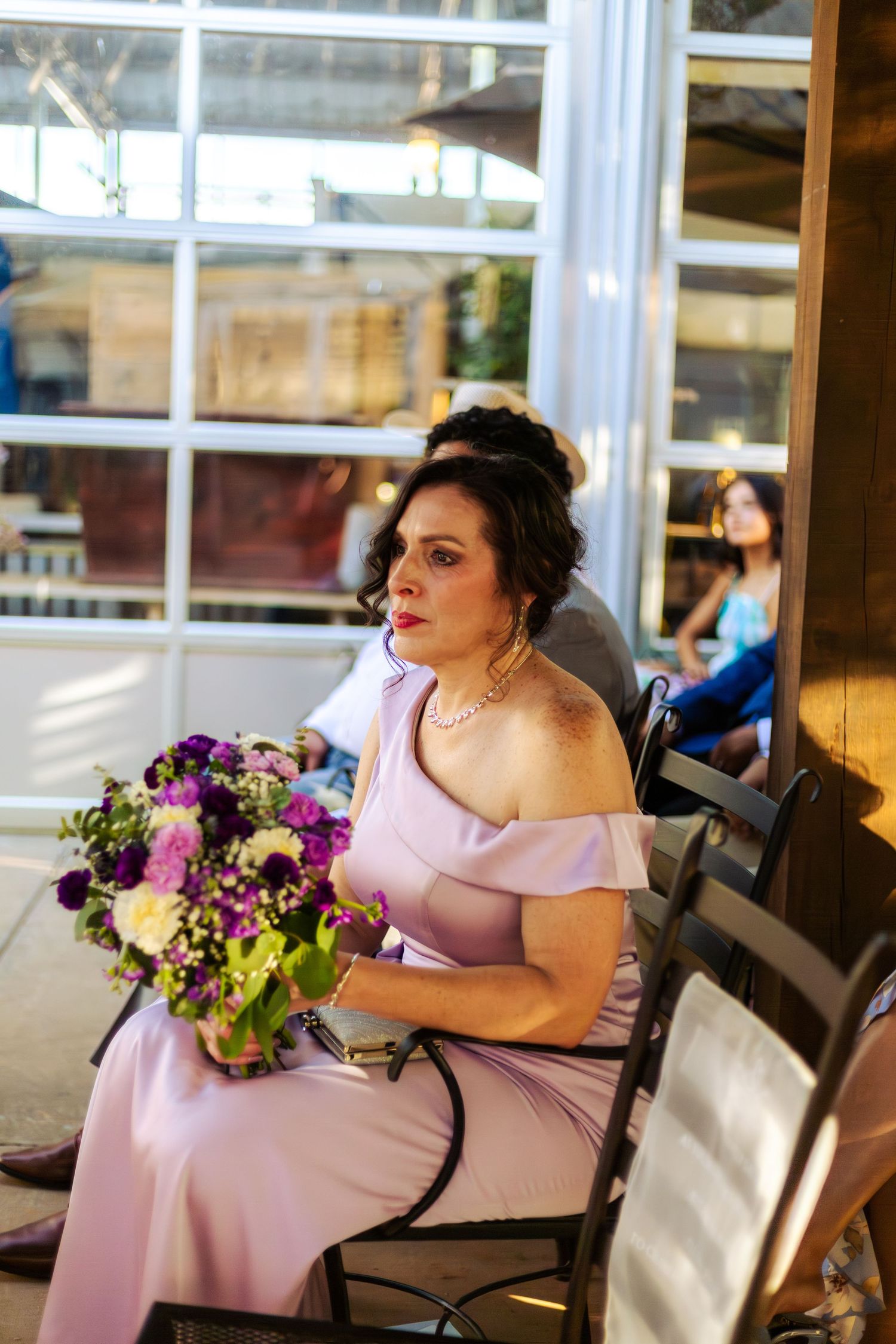Woman in lavender off-shoulder gown holding purple floral bouquet seated at indoor wedding venue.