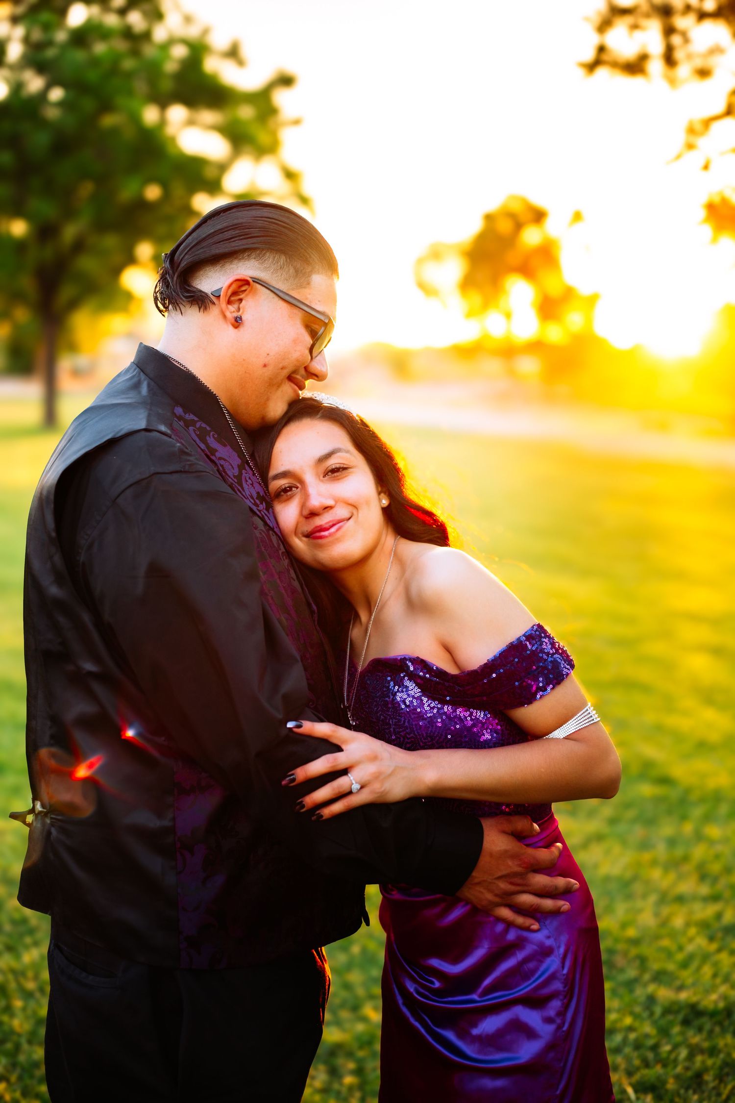 Couple embracing at golden hour sunset in a park, woman in purple sequin gown, man in black suit with glasses.