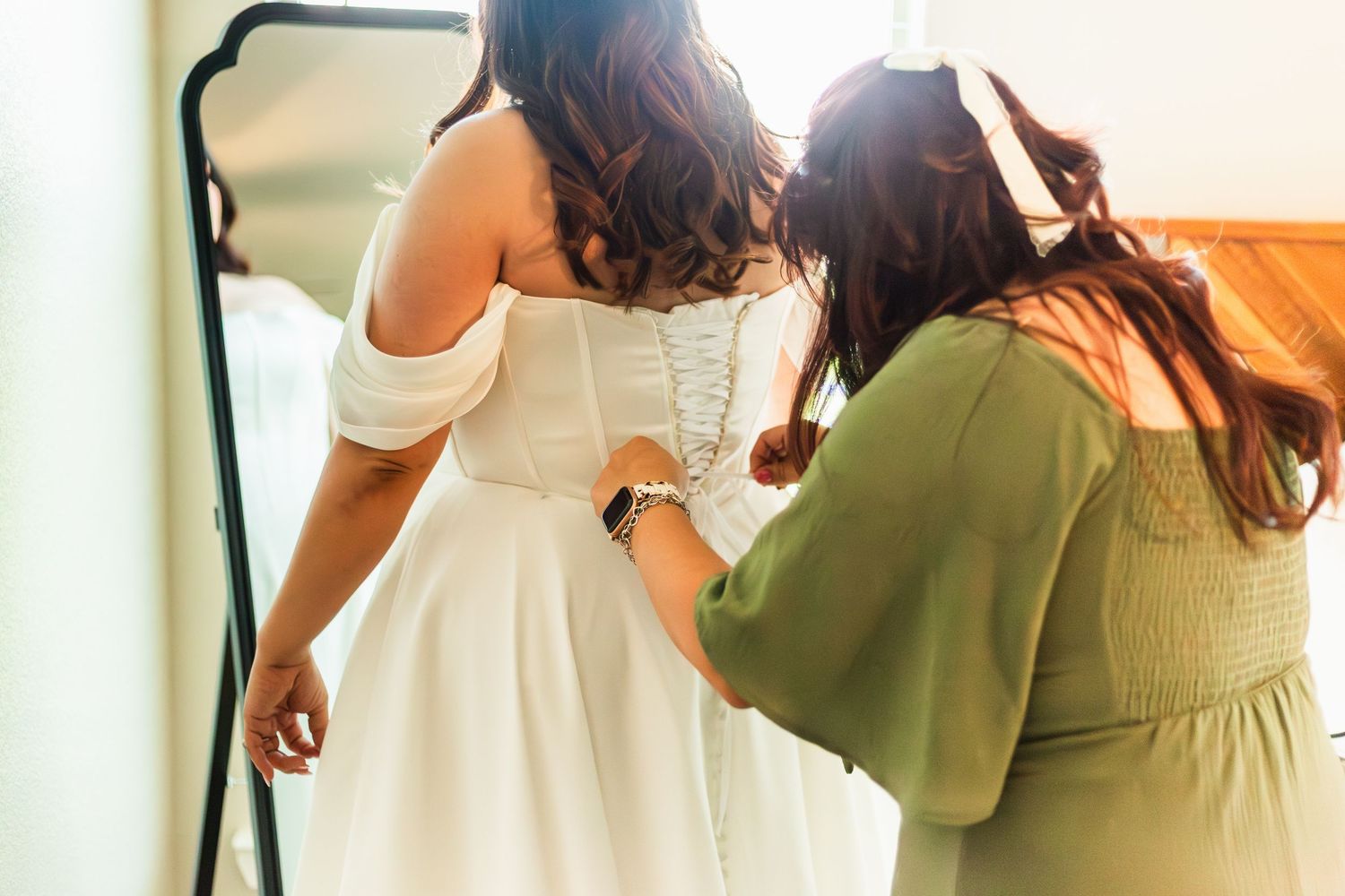 A bridesmaid in green helps a bride button her white off-shoulder wedding dress in front of a mirror.