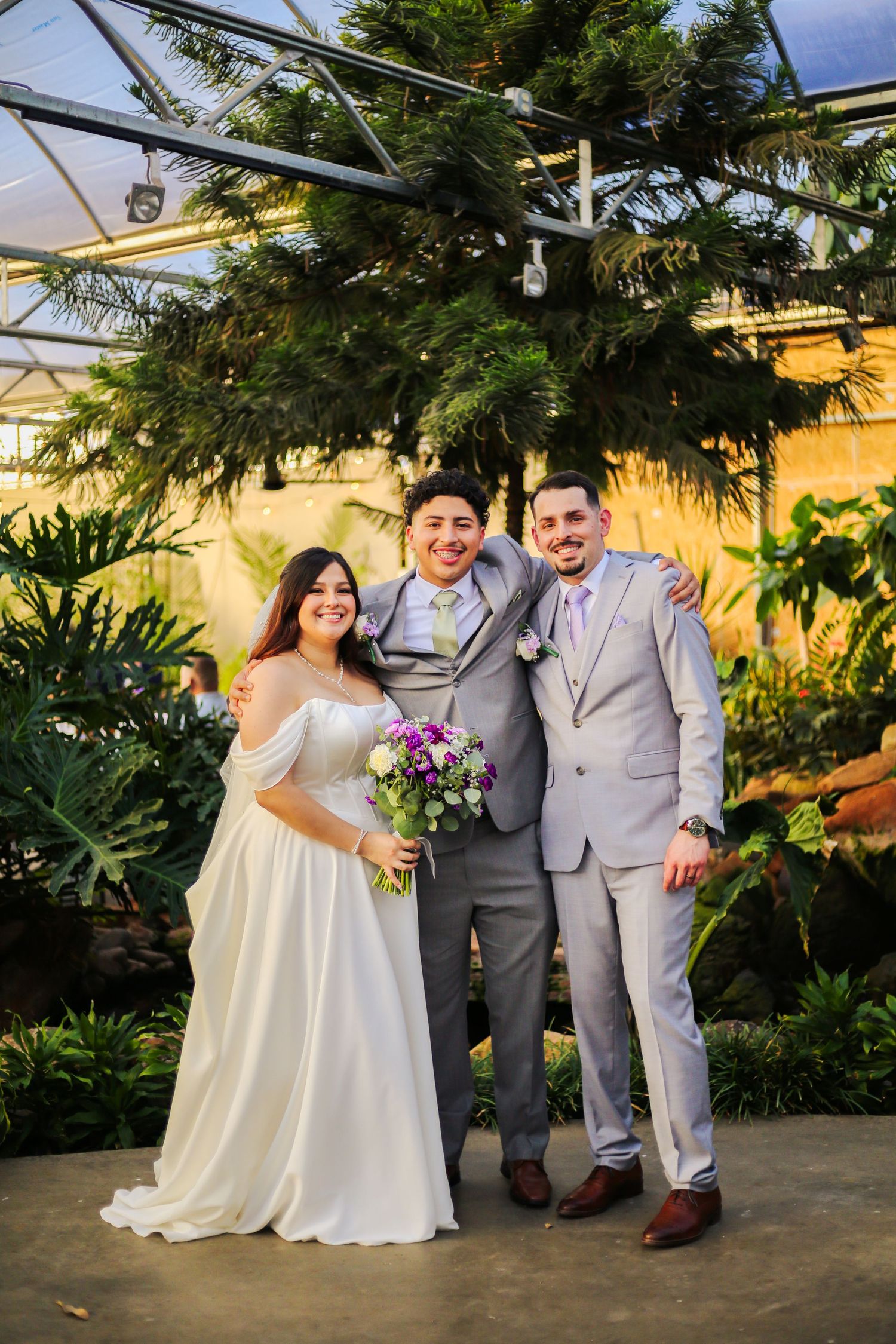 Bride and groom pose with groomsman in gray suits inside a lush greenhouse venue holding purple wedding bouquet.