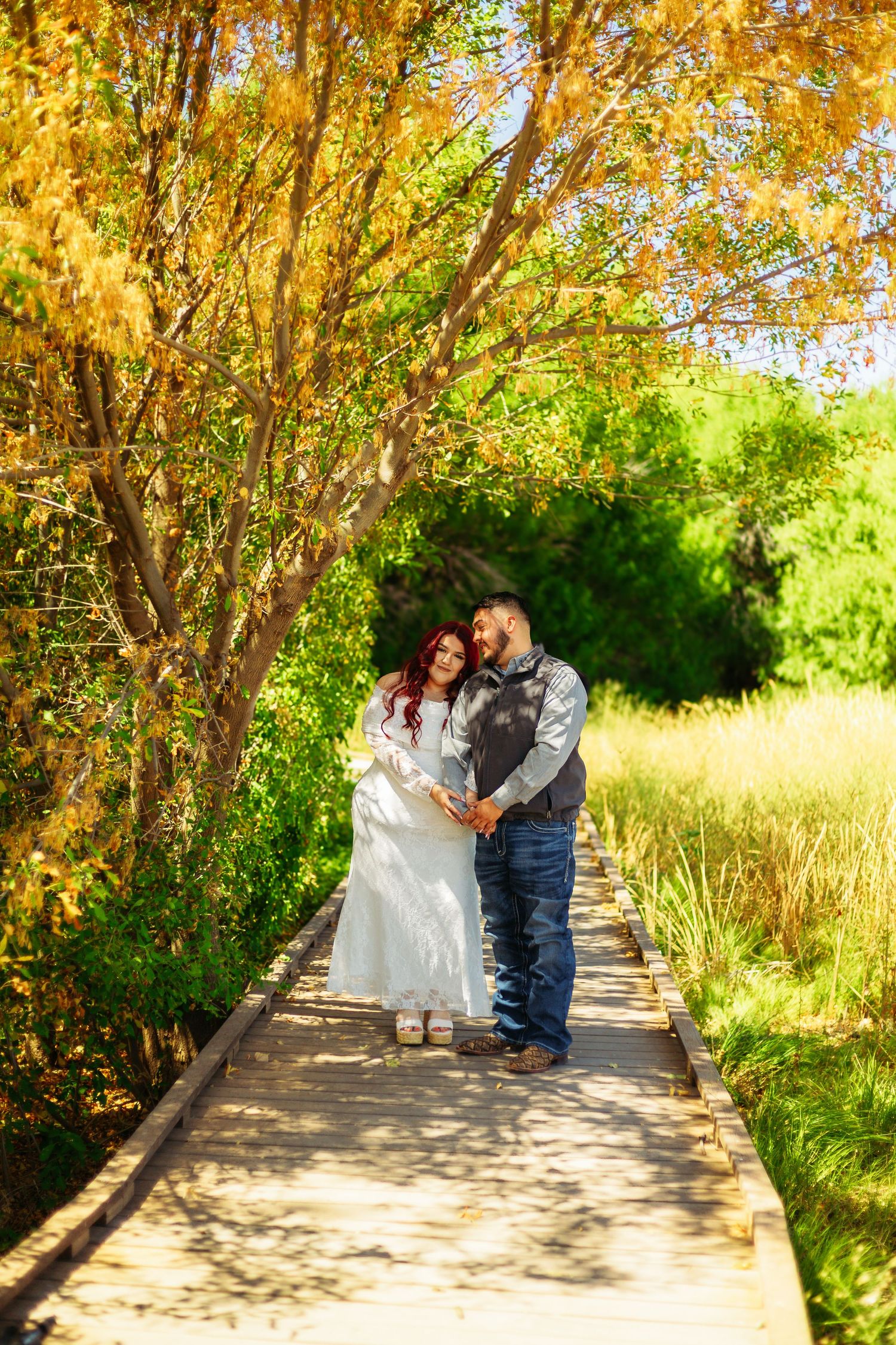 Couple holding hands on wooden boardwalk path surrounded by autumn yellow and green trees in a romantic outdoor setting.