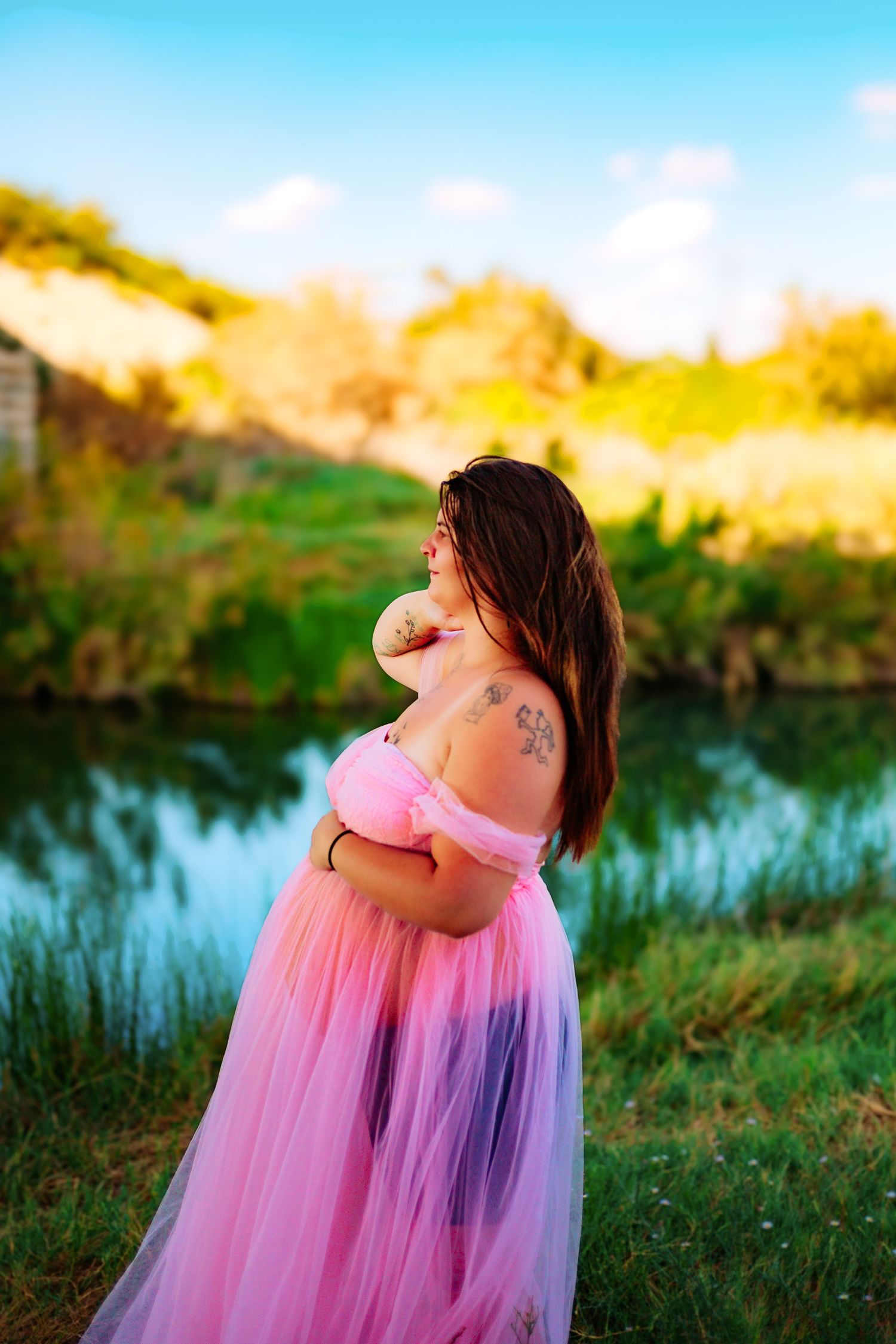 Pregnant woman in pink tulle gown poses outdoors near a reflective pond surrounded by lush greenery under a blue sky.